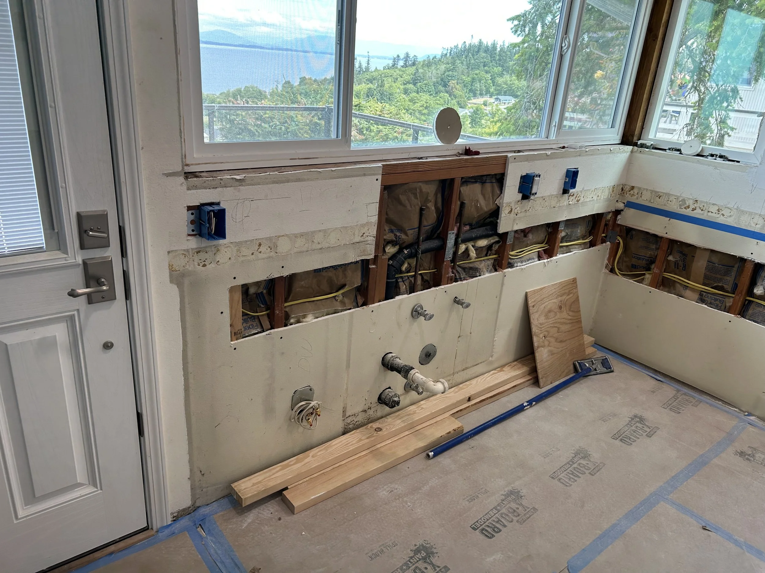 Kitchen renovation with open wall showing plumbing and electrical work, construction materials on the floor, and large windows with scenic outdoor view.