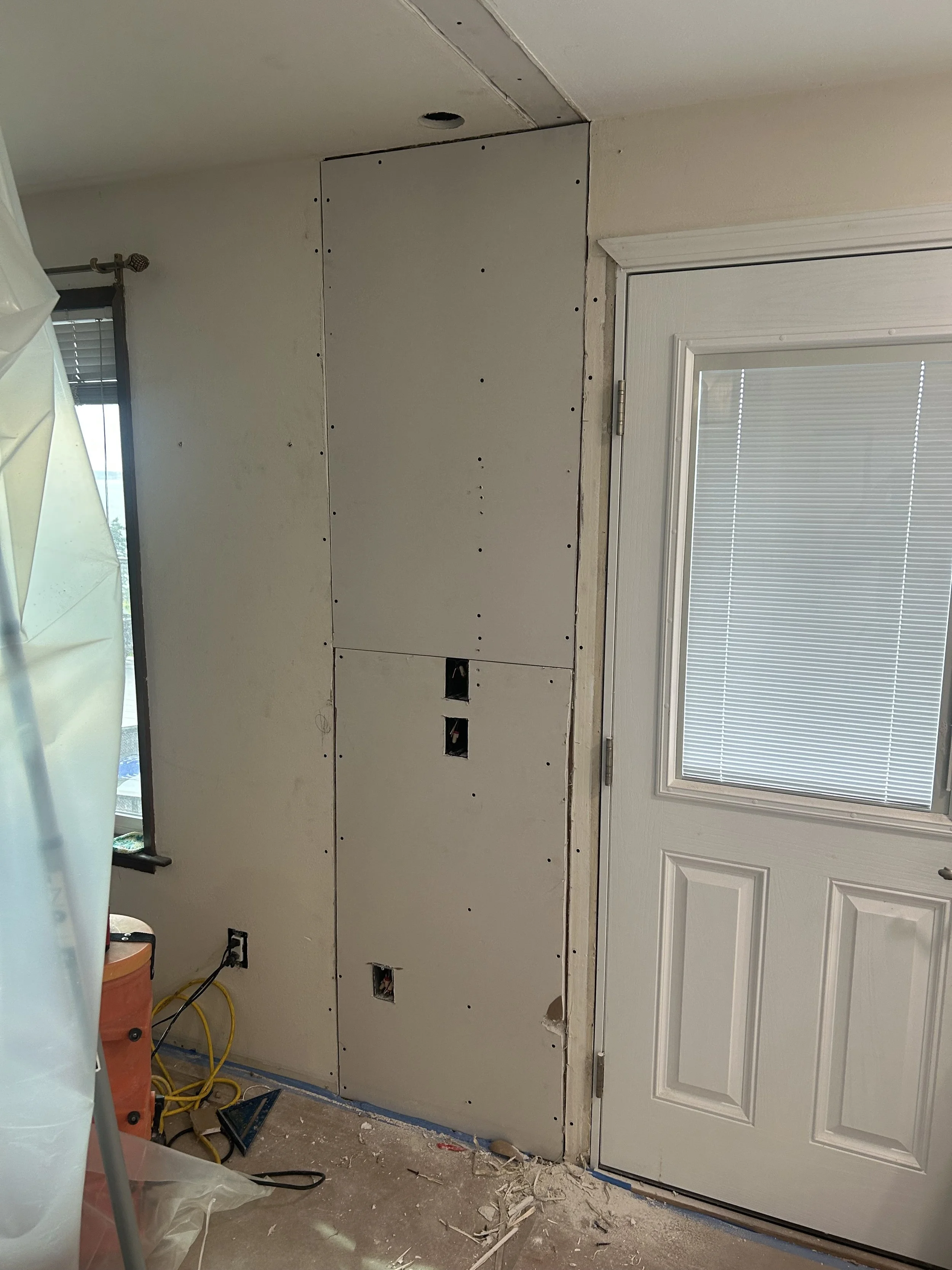 Interior wall under renovation with drywall panels, electrical outlets, and wiring, next to a door with a window