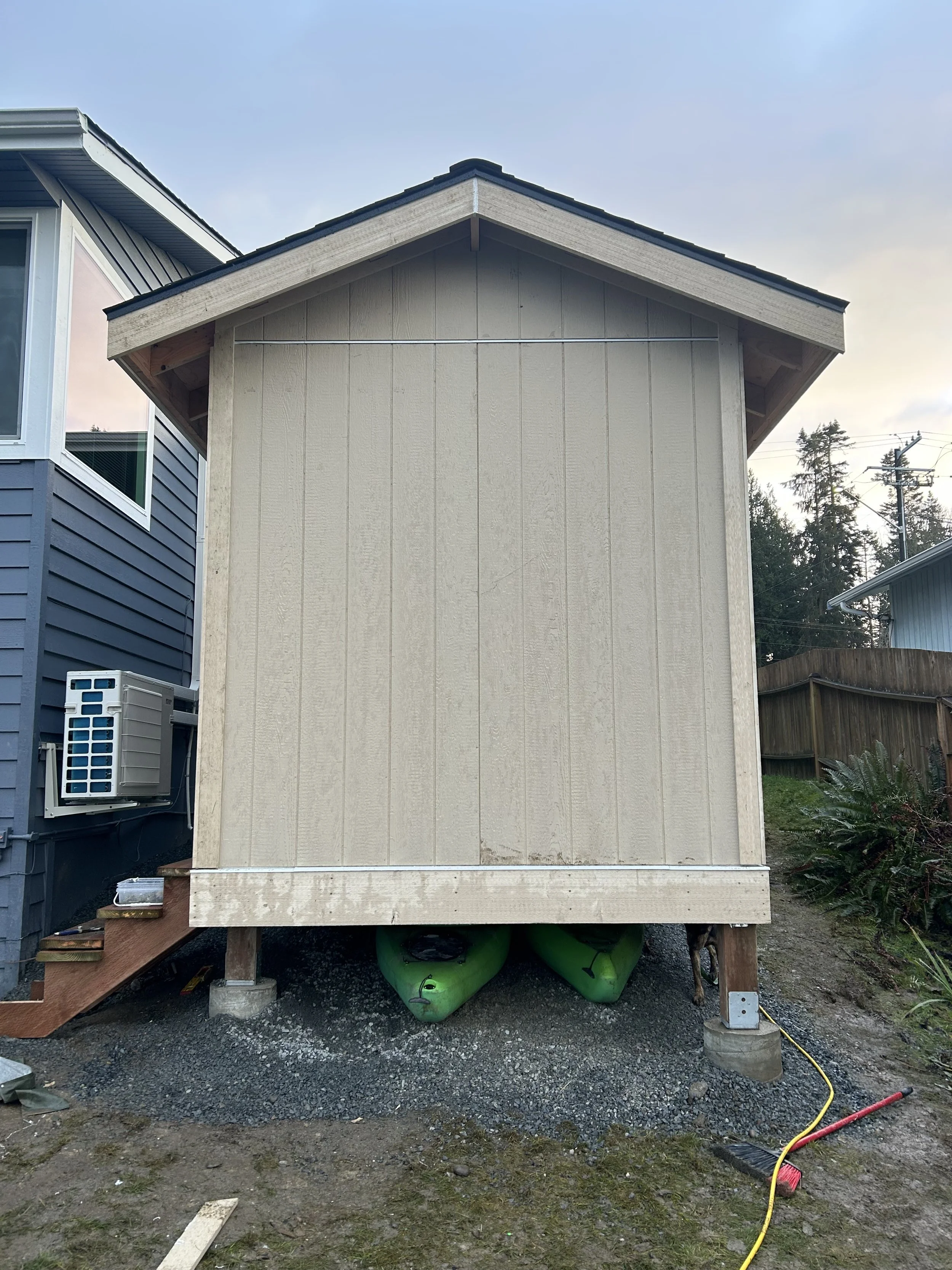 Small beige shed on stilts with a sloped roof, two green kayaks stored underneath, and construction materials and tools nearby.
