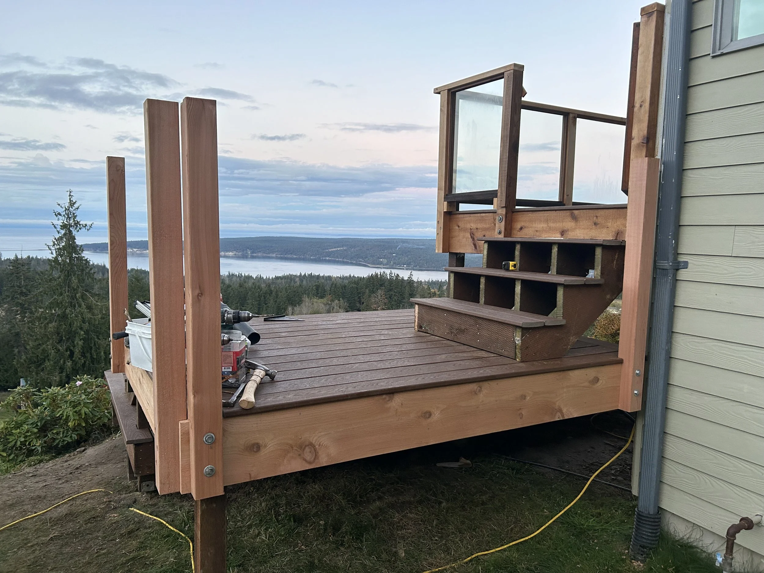 Wooden deck under construction beside a green house, with tools and construction materials, overlooking a lake and forested landscape.