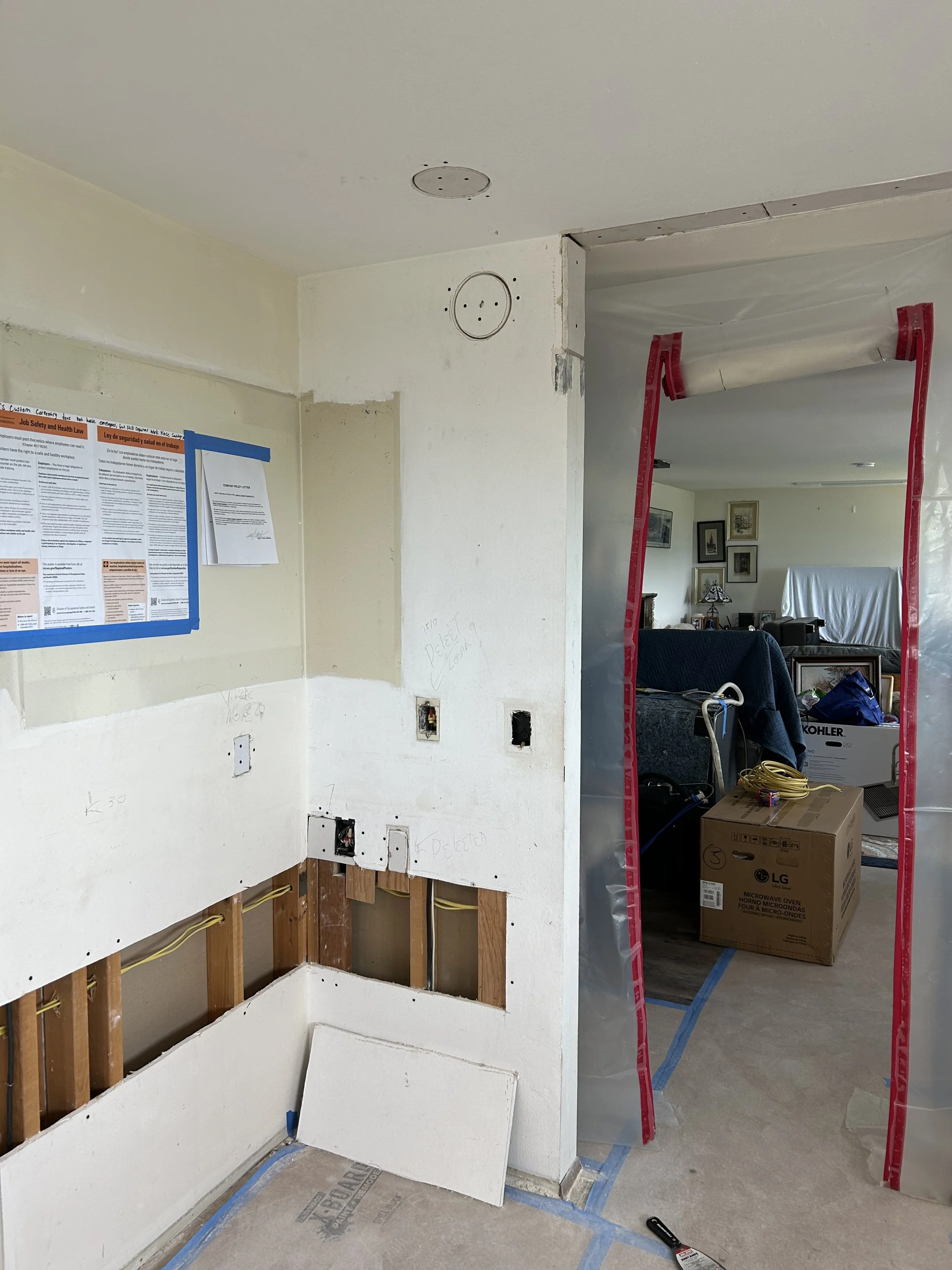 Room under renovation with exposed wall studs, electrical wiring, and partially installed ceiling fixtures. There is a doorway taped off with red protective film, and a living room with furniture and boxes is visible in the background.