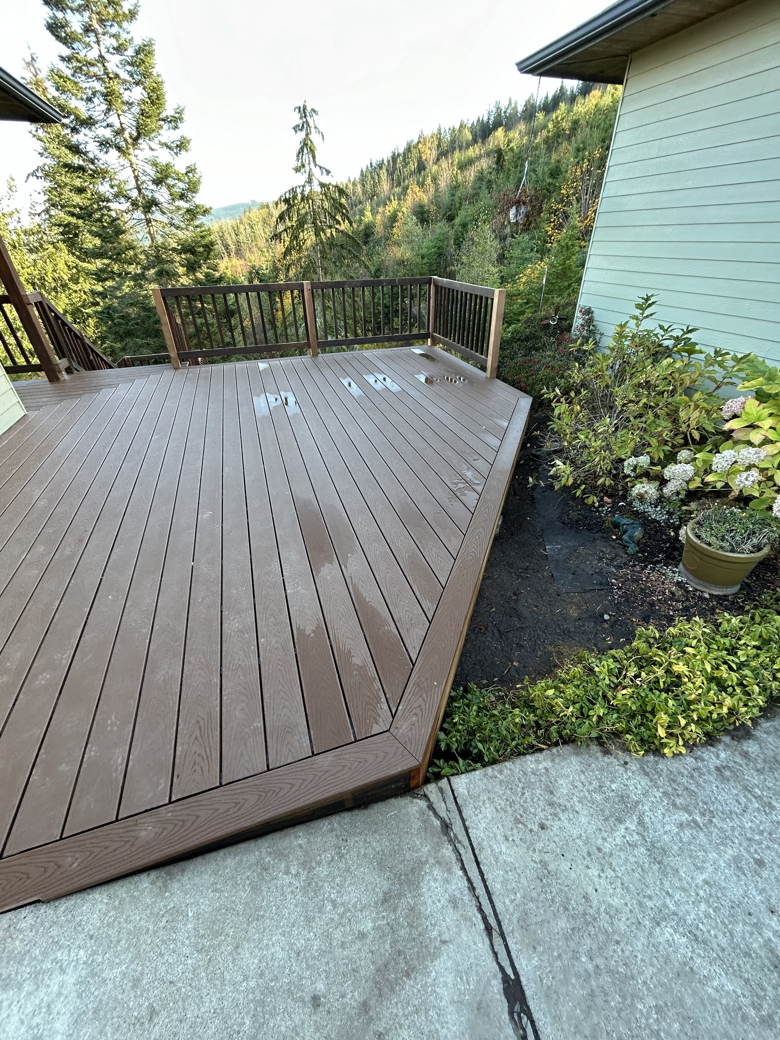 An outdoor wooden deck attached to a house with a view of green trees and forested hills in the background. The deck has a railing on one edge and is adjacent to a garden bed with various plants and flowers.