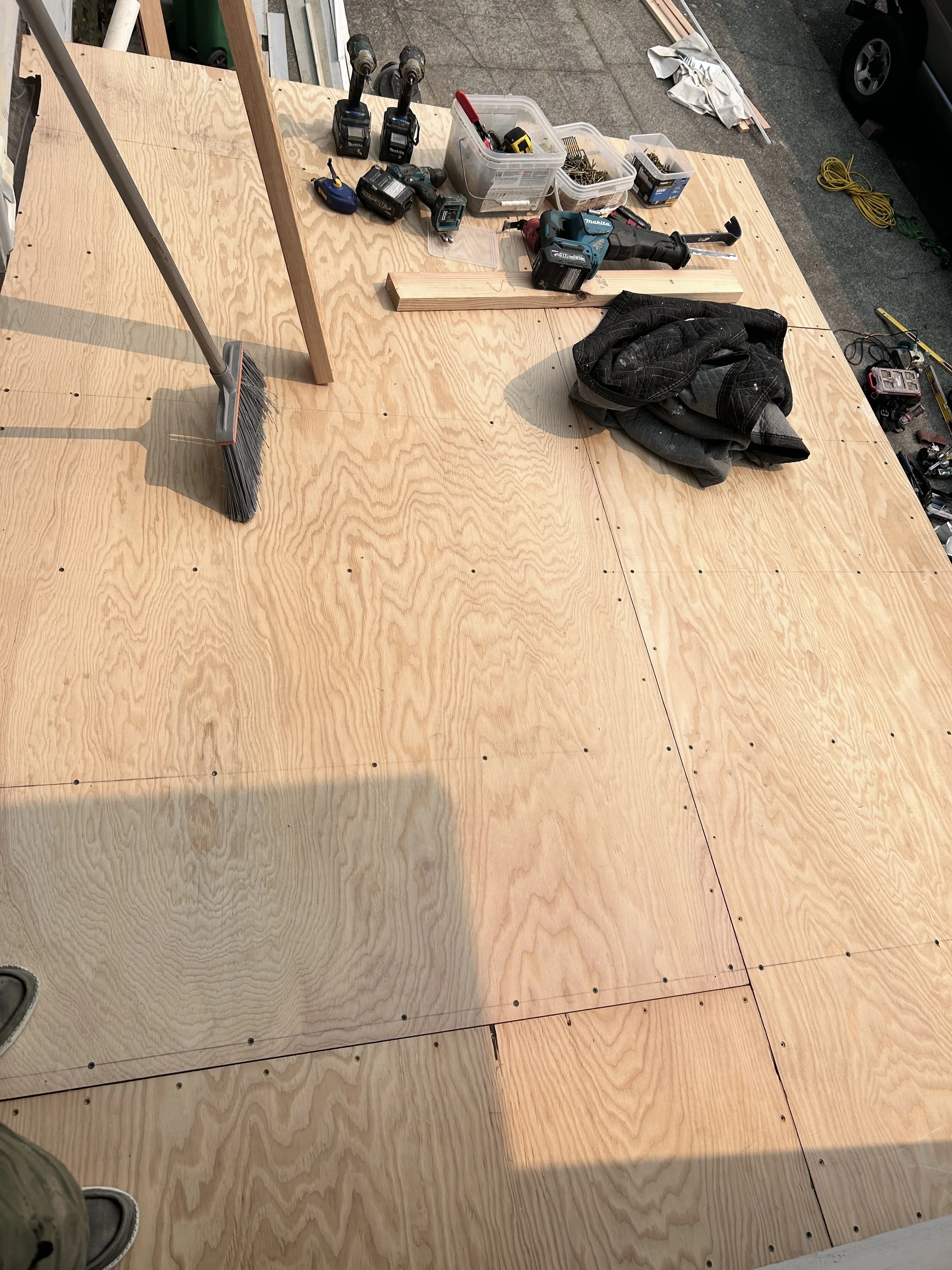 A construction workspace with plywood sheets, tools including drills, a saw, a broom, and a toolbox, and a black jacket on the plywood surface.