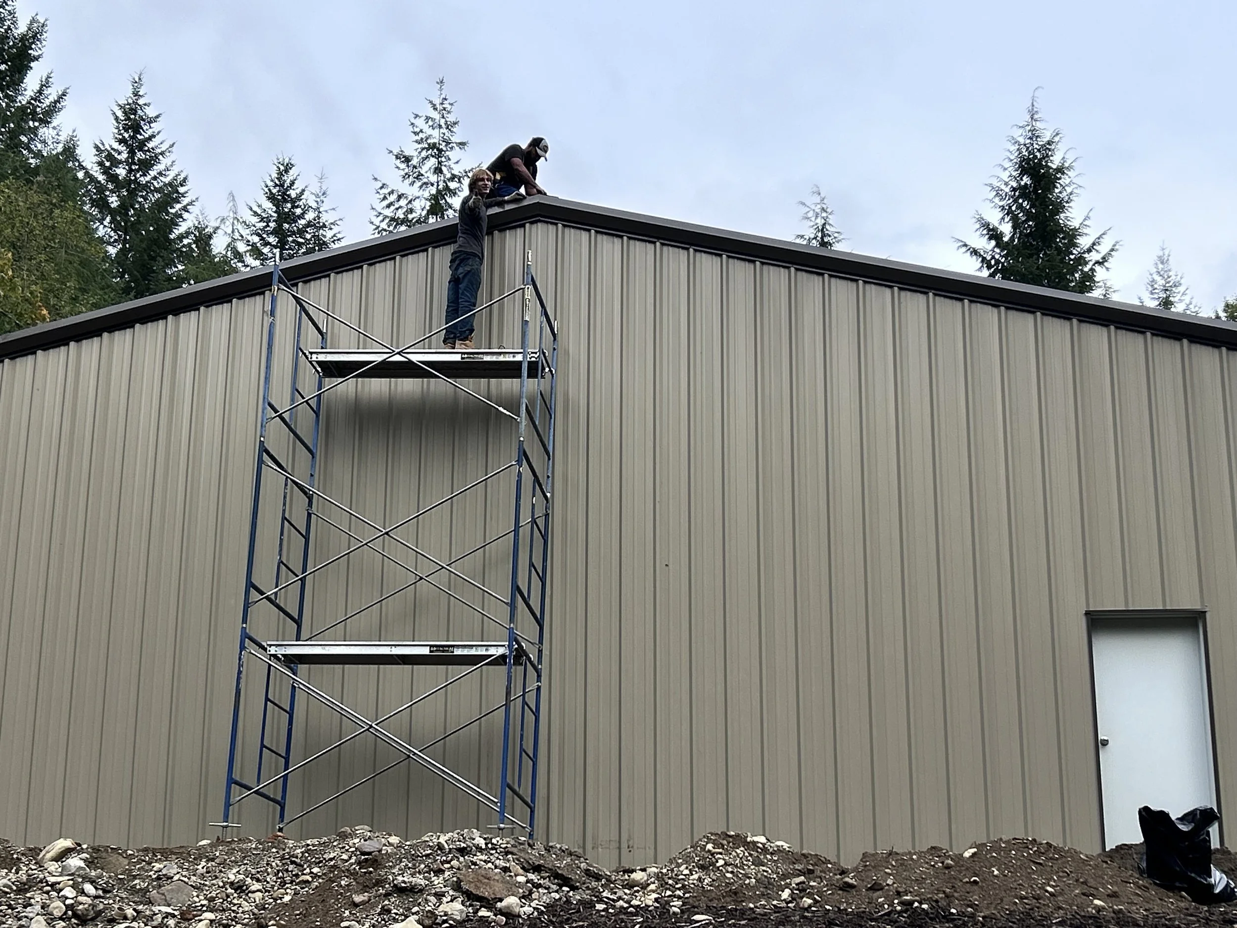 Two workers on a scaffold installing or maintaining a roof on a large metal building, with trees in the background.