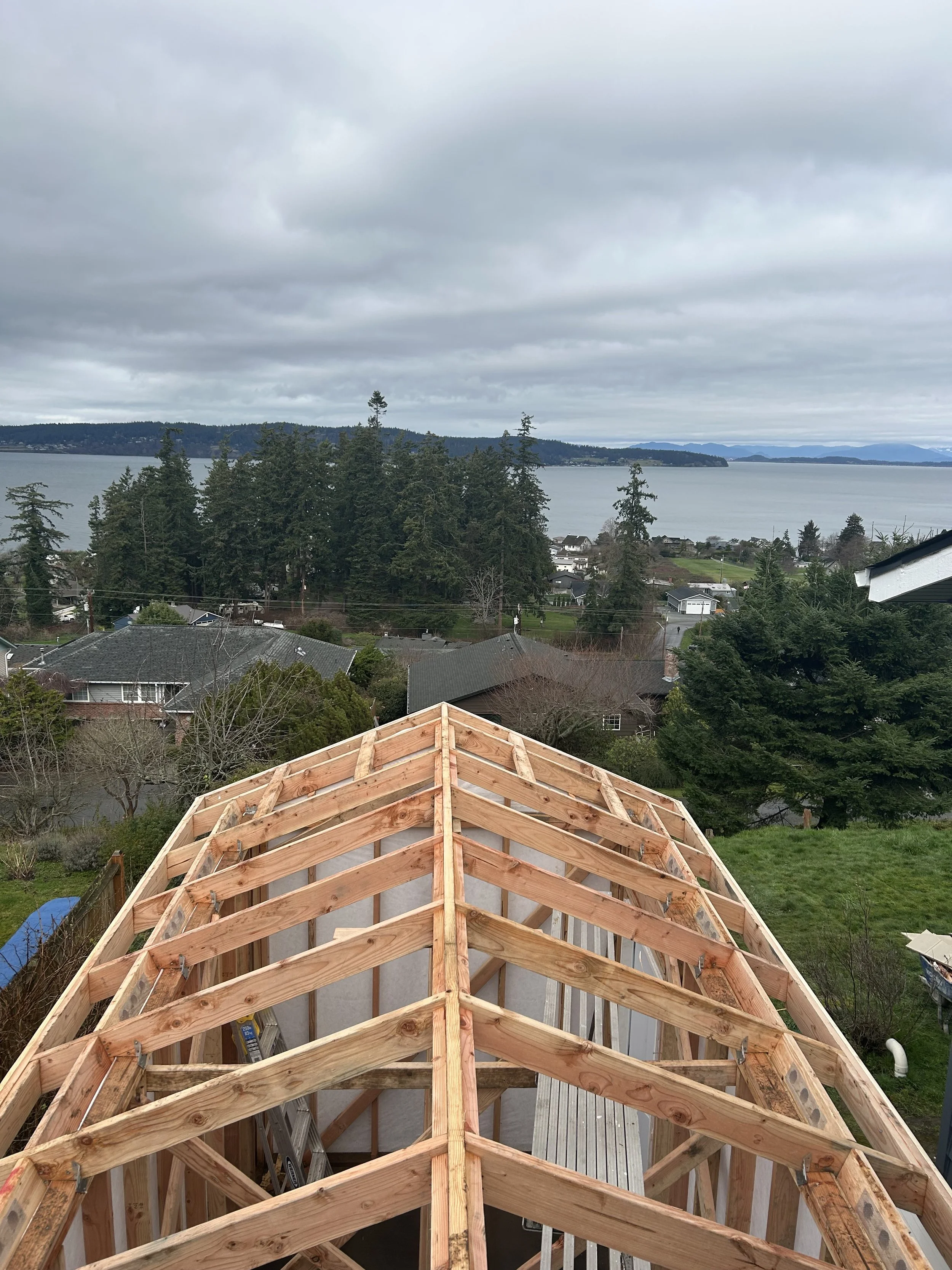 Construction of a roof with exposed wooden beams and a view of a residential neighborhood, trees, and a body of water under a cloudy sky.