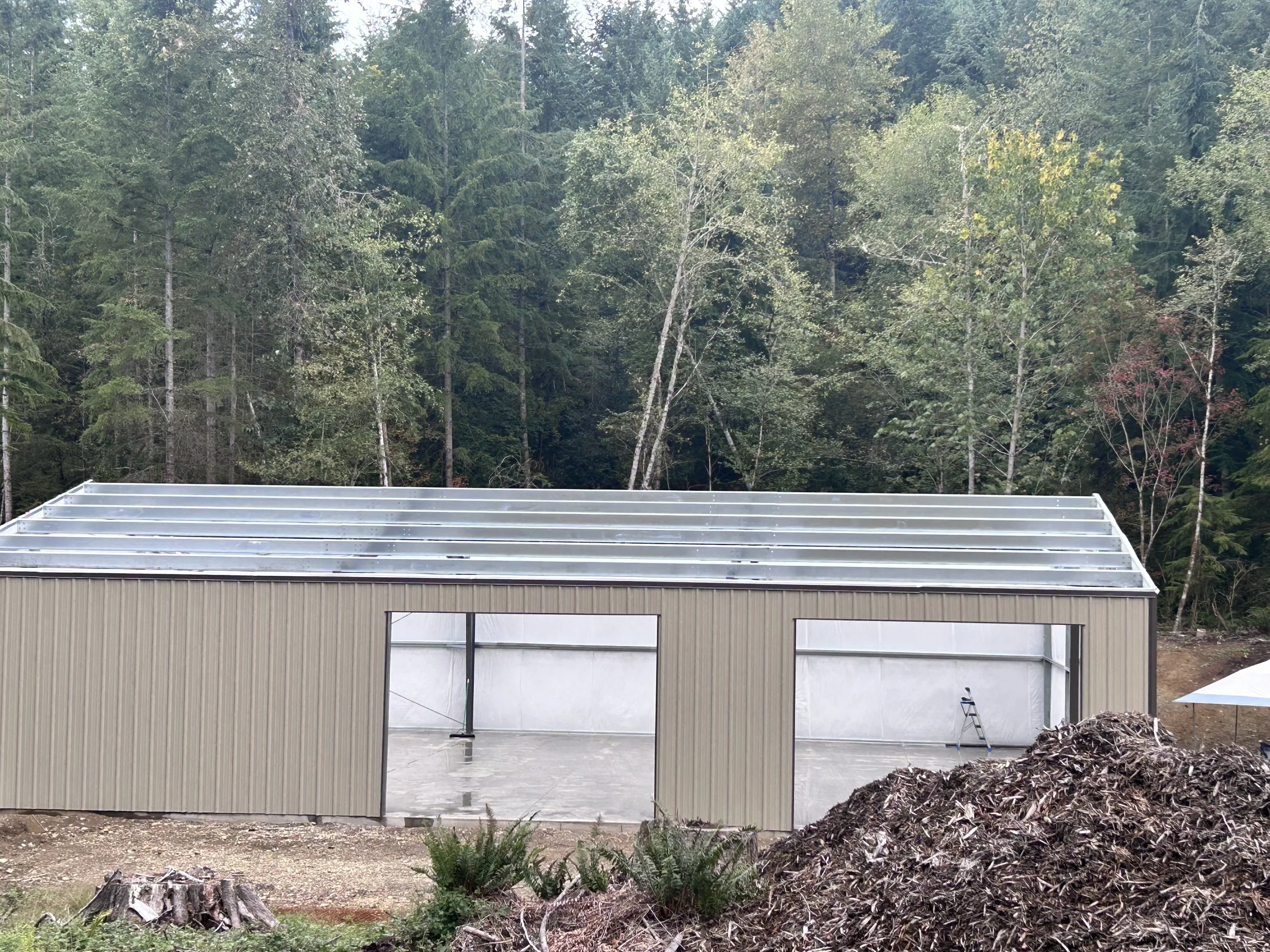 Under construction metal building with open garage doors, set against a forested background with trees.