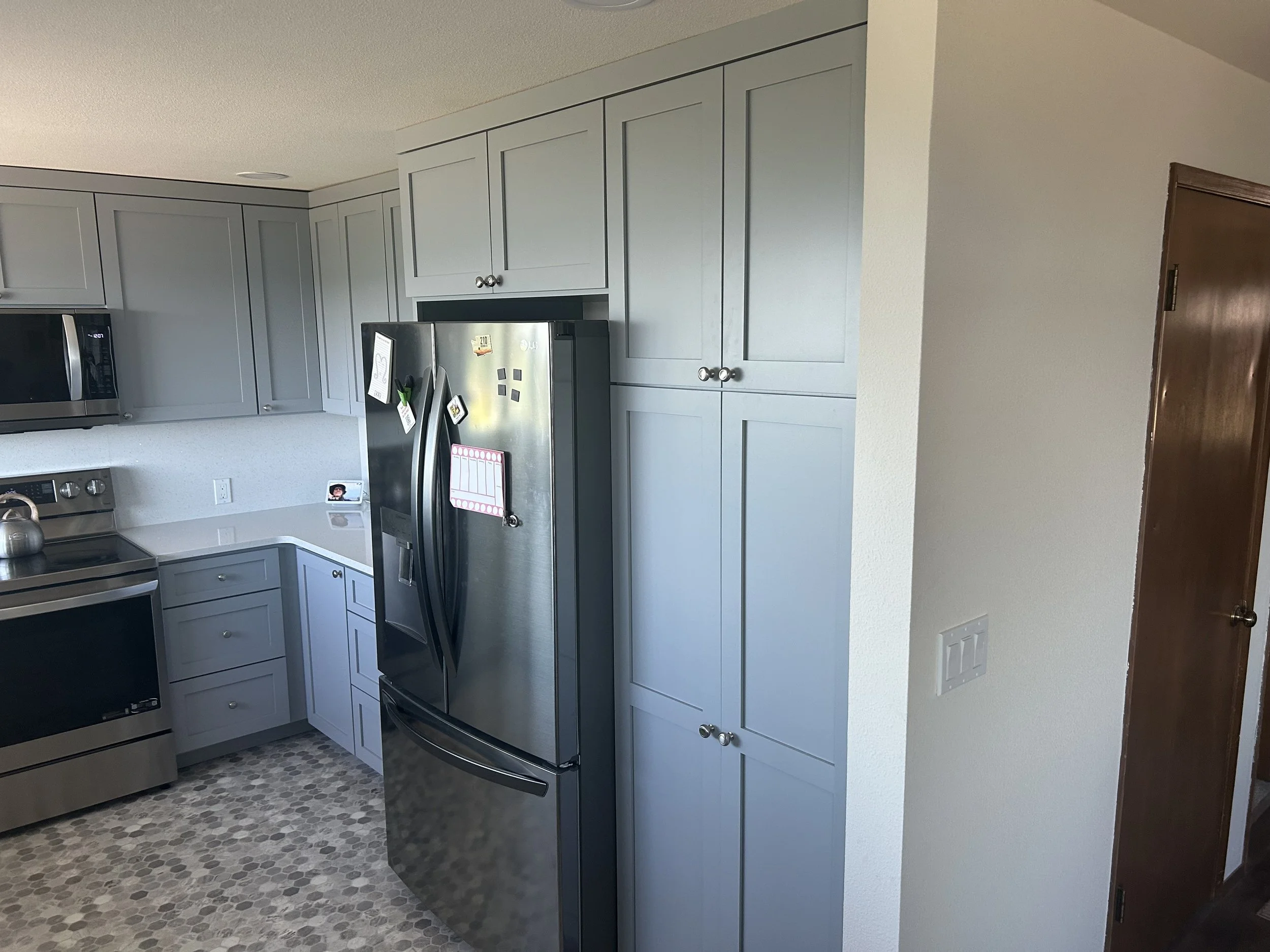 Modern kitchen with gray cabinets, stainless steel refrigerator with magnets and notes, stove, microwave, and patterned tile floor.