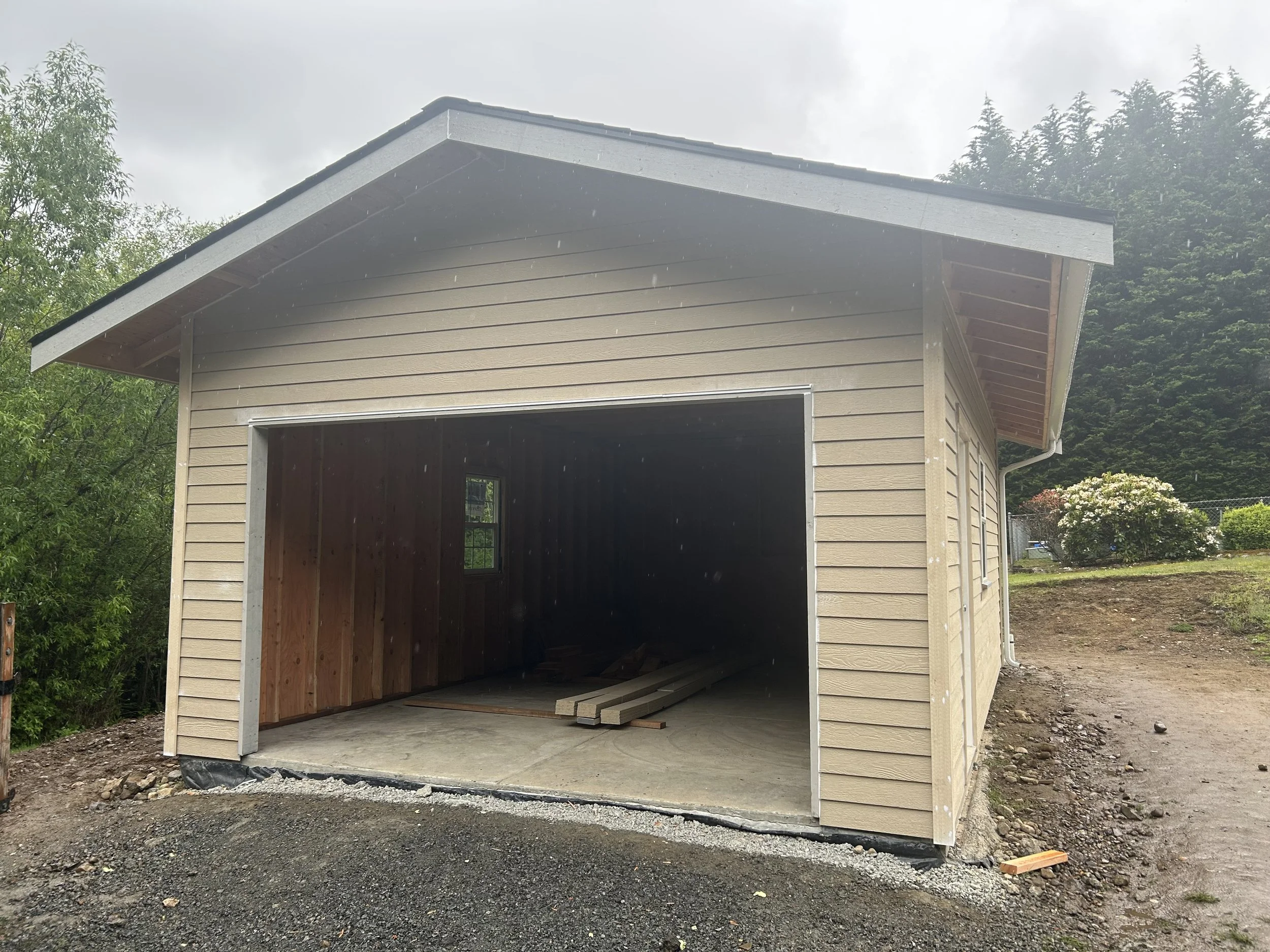 A partially built garage with beige siding and a dark brown interior, construction materials inside, on a gravel driveway with greenery and trees around.