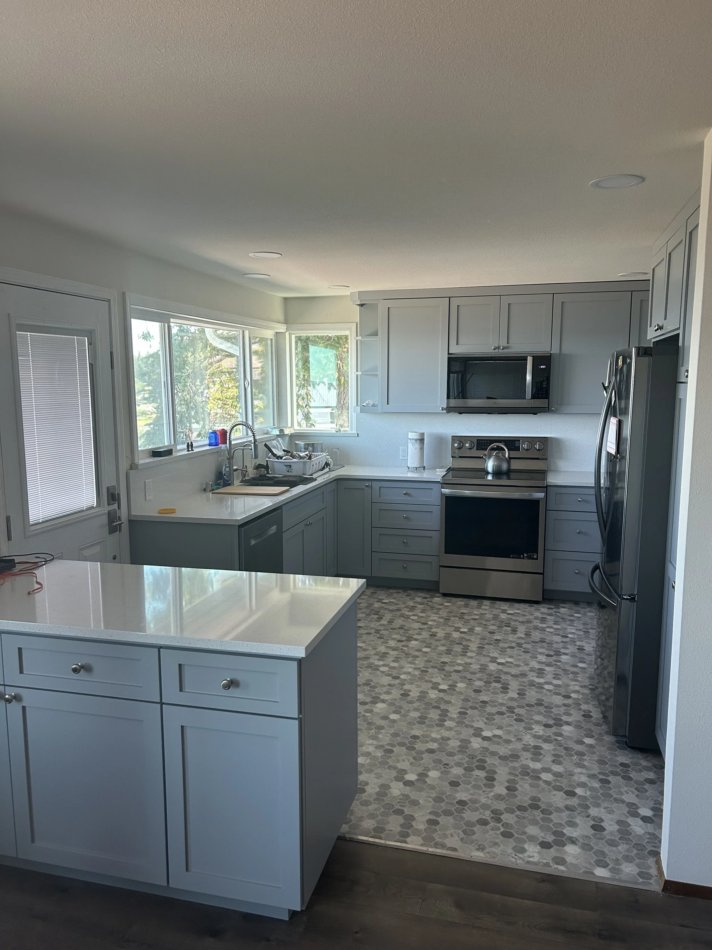 Modern kitchen with gray cabinets, stainless steel appliances, and a breakfast island. Sunlight streams through large windows.