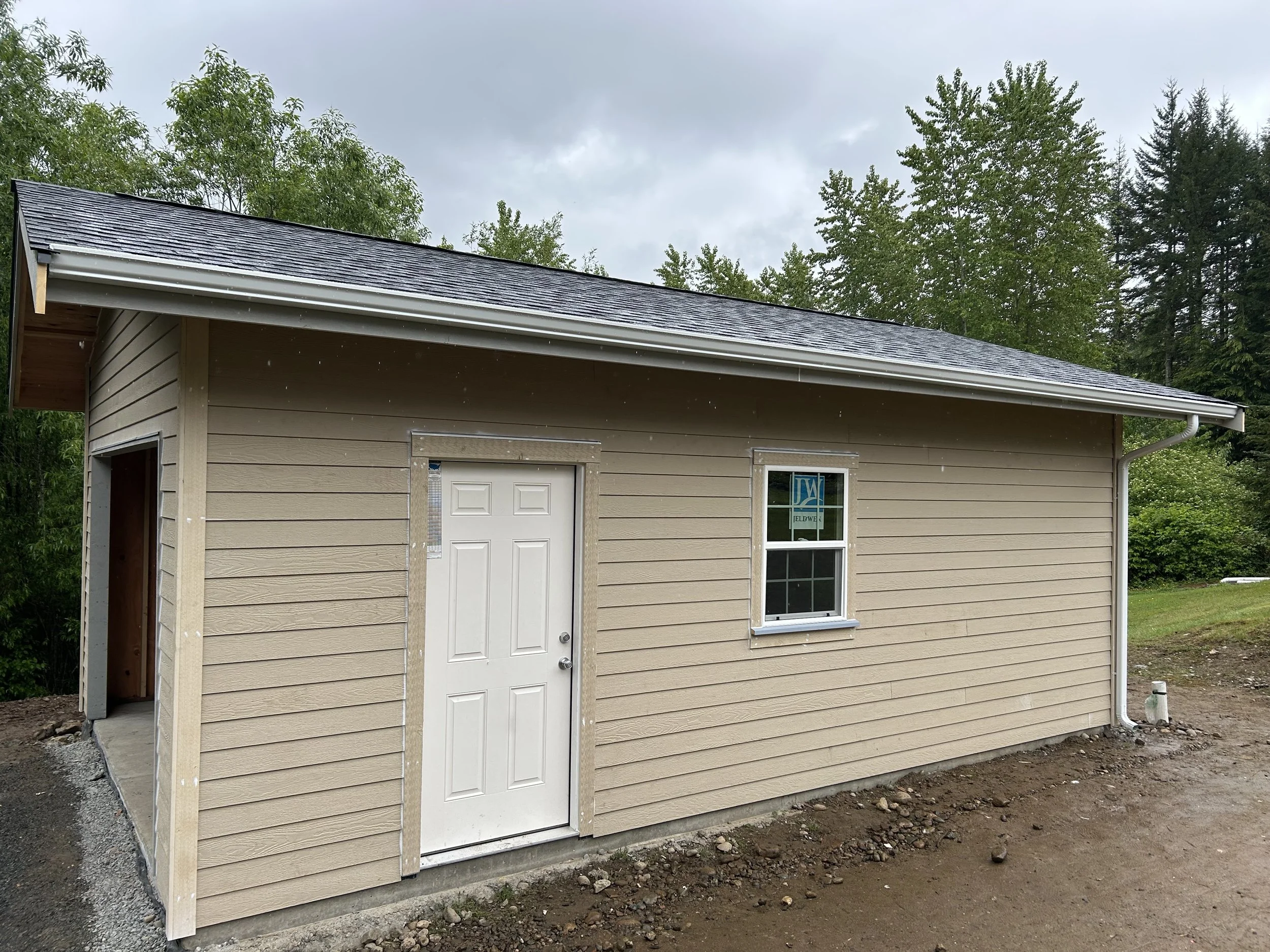 New beige house with a single door and window, surrounded by a dirt yard and green trees under an overcast sky.