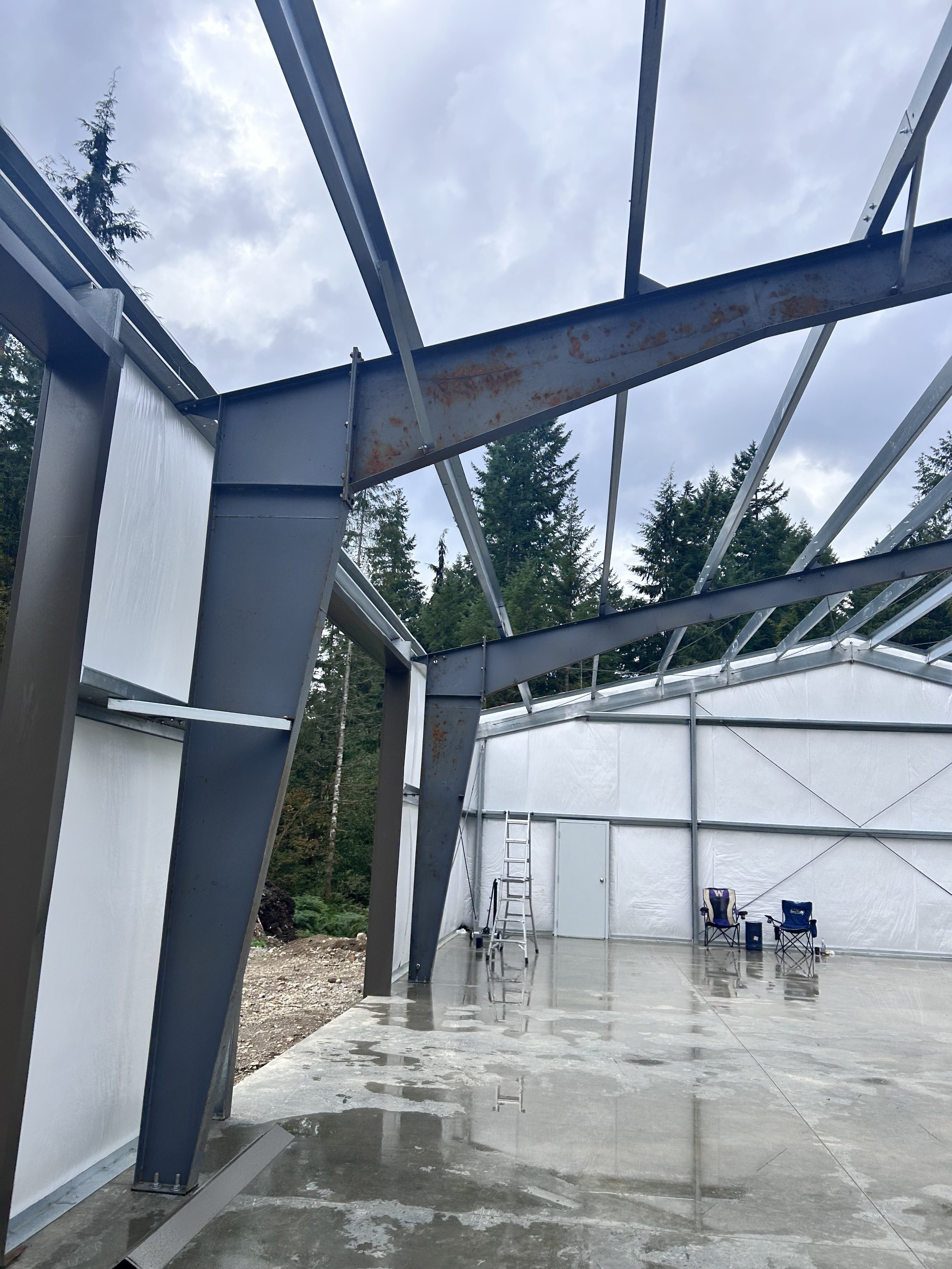 Interior of a partially constructed building with exposed metal framing, a ladder, two chairs, and a wet concrete floor, surrounded by green trees and cloudy sky.