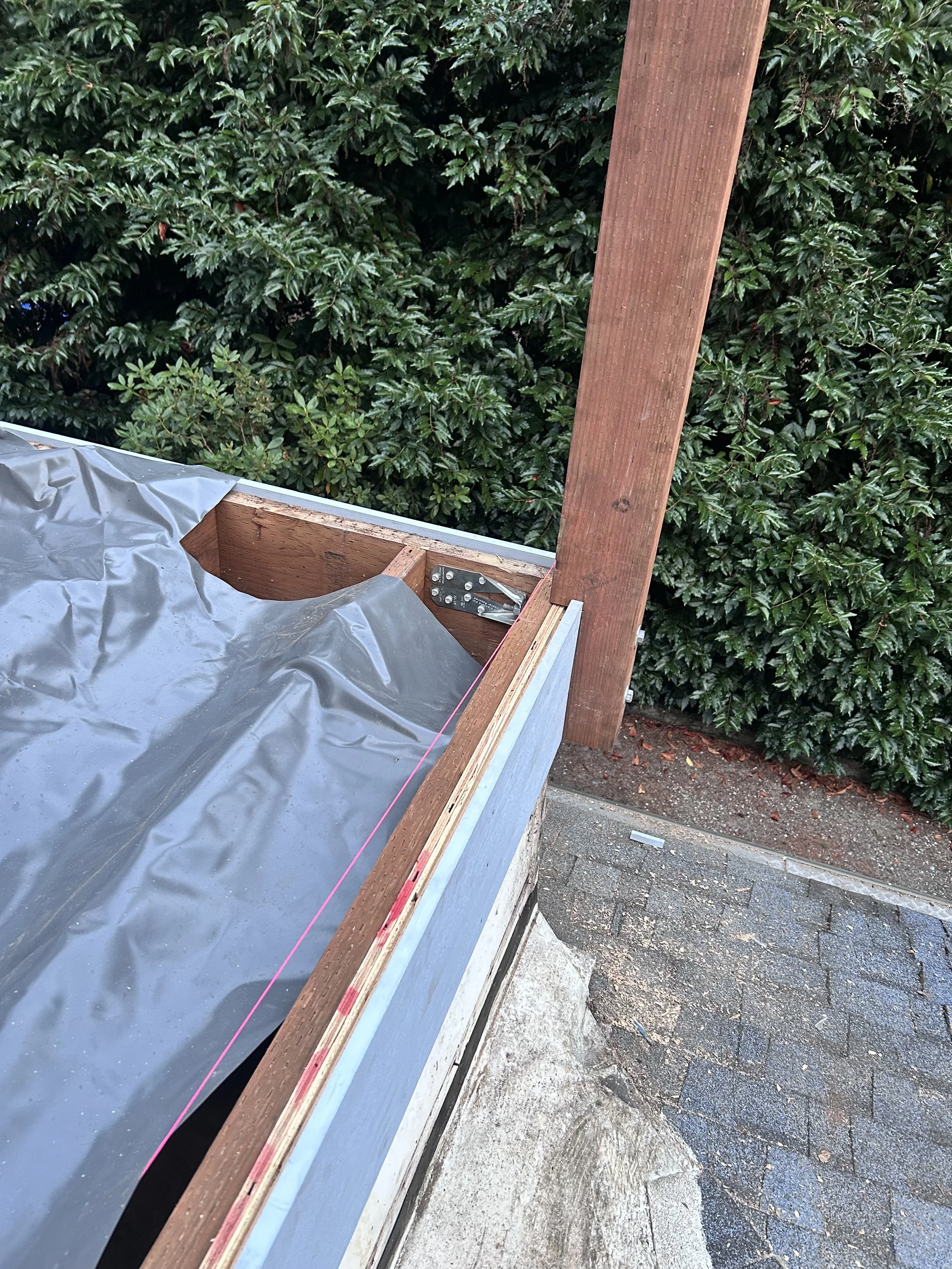 Roof with underlayment and wooden framing, overlooking a hedge and brick pavement.