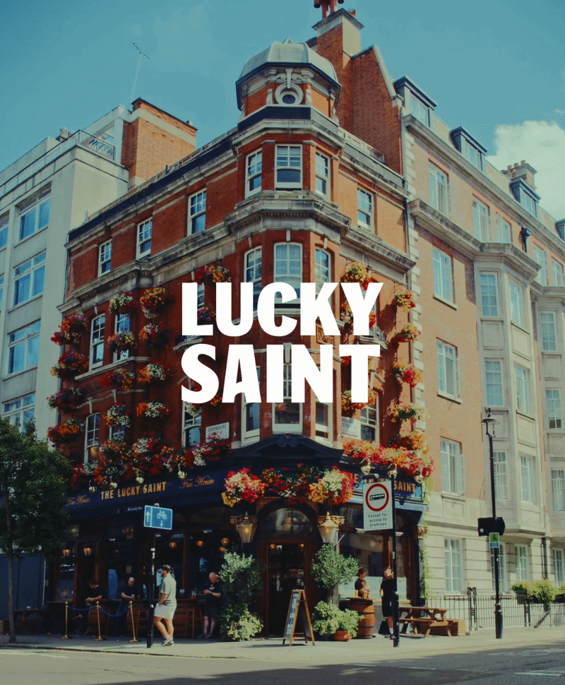 A multi-story corner building with a pub named 'The Lucky Saint' on the ground floor, decorated with colorful flower baskets, with people walking by on a sunny street, under a clear blue sky.