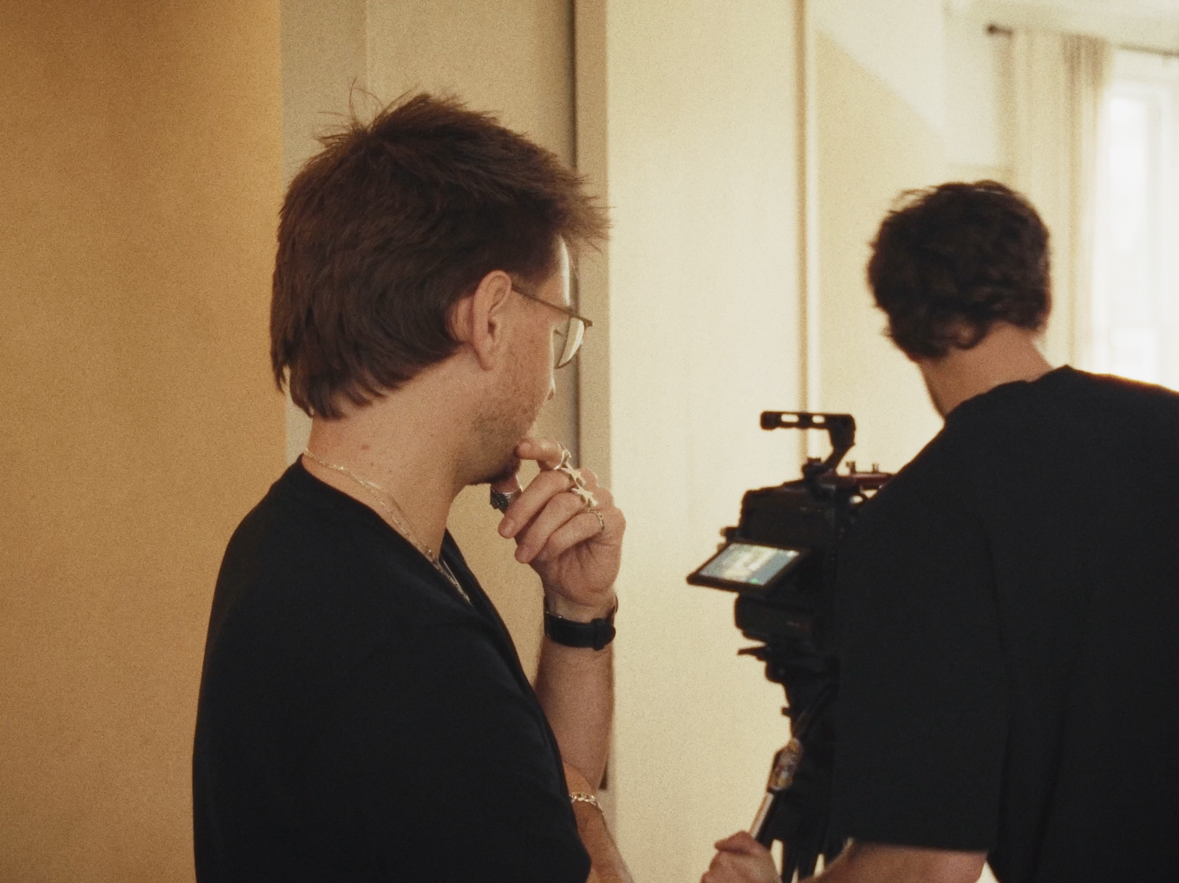 Two people, one with long brown hair and glasses, holding a camera, looking at it while standing indoors near a window with curtains.