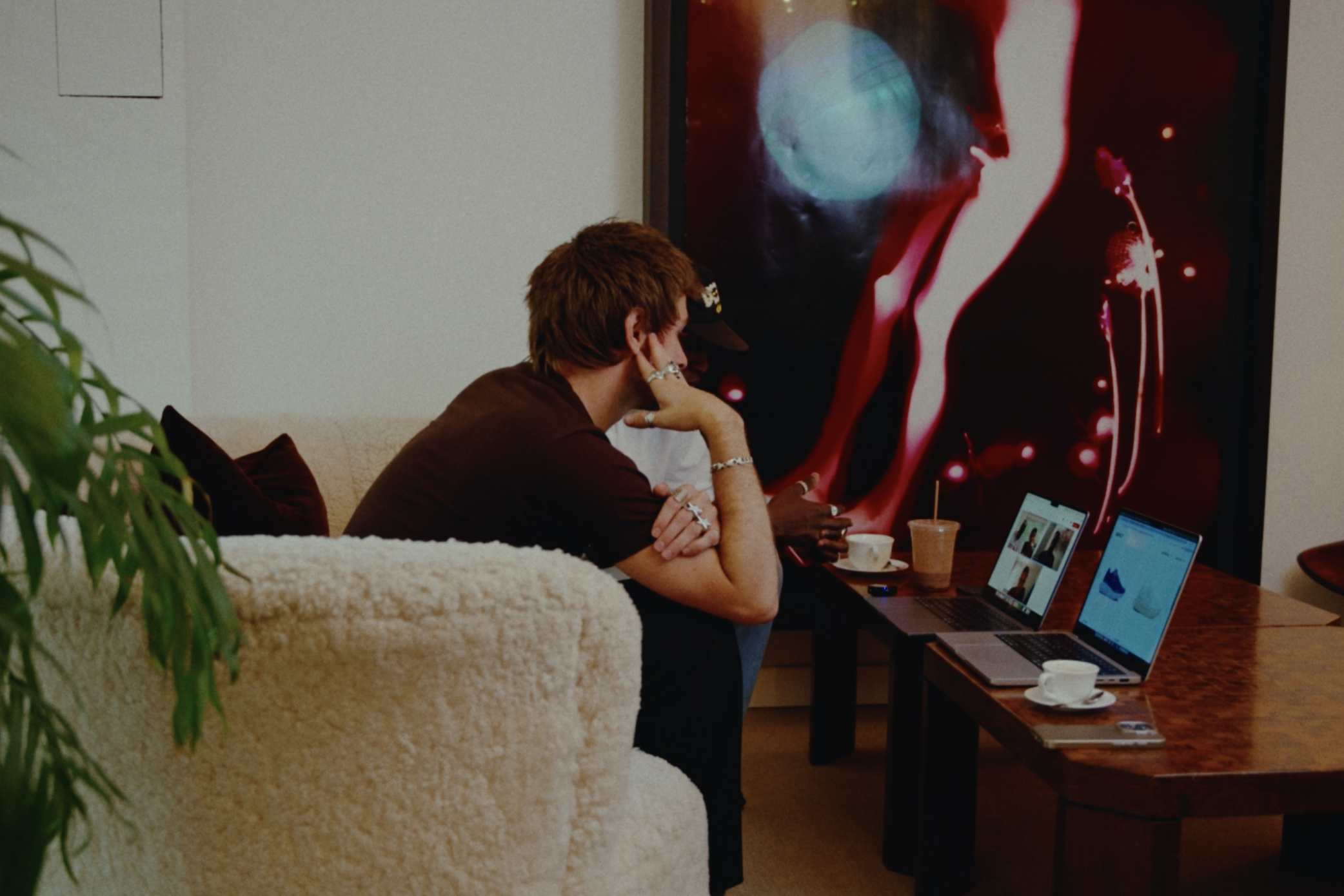 A man with short brown hair, sitting on a beige sofa, looking at multiple laptops on a wooden table, with drinks and cups, in front of an abstract cityscape poster on the wall.
