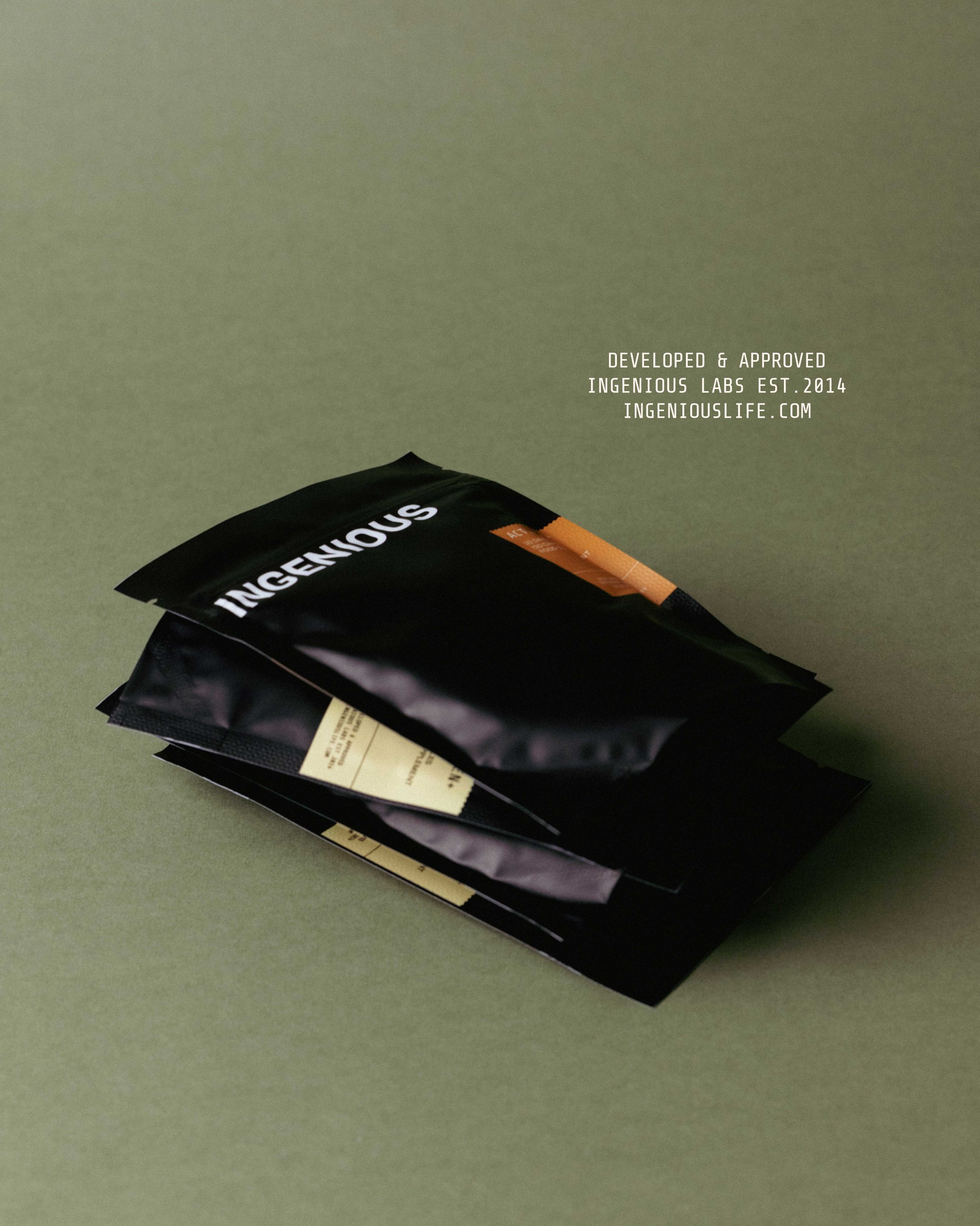 Stack of black packaging pouches on a green surface with white text about Ingenious wellness company.