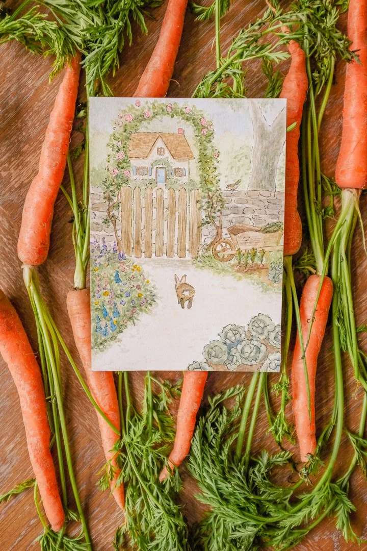 Watercolor painting of a bunny hopping on a path towards a garden gate with a cottage in the background