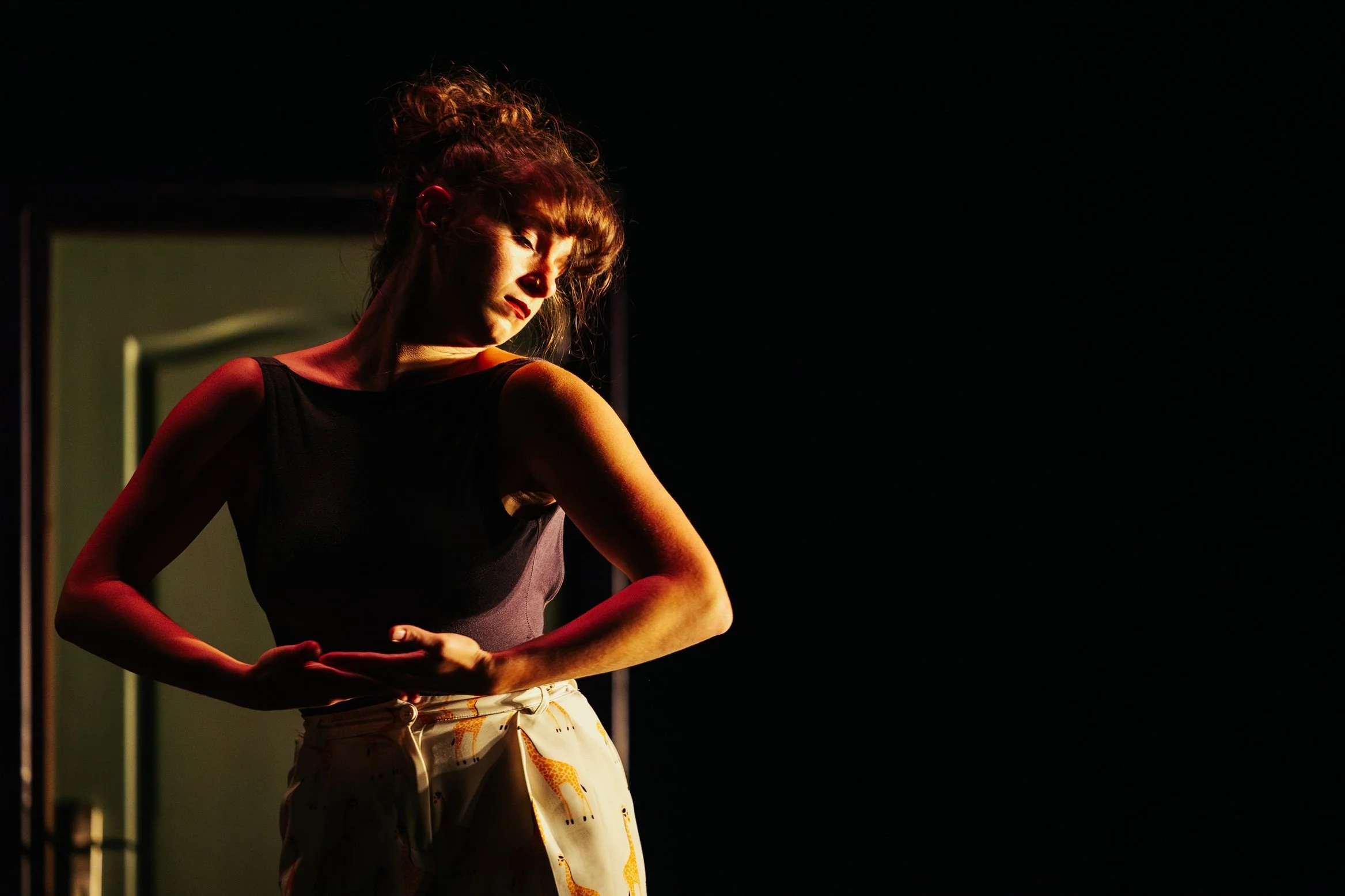 Femme avec cheveux bouclés dans une expression concentrée, habillée d’un haut noir et de pantalons à motif girafe, dans une lumière chaude et sombre.