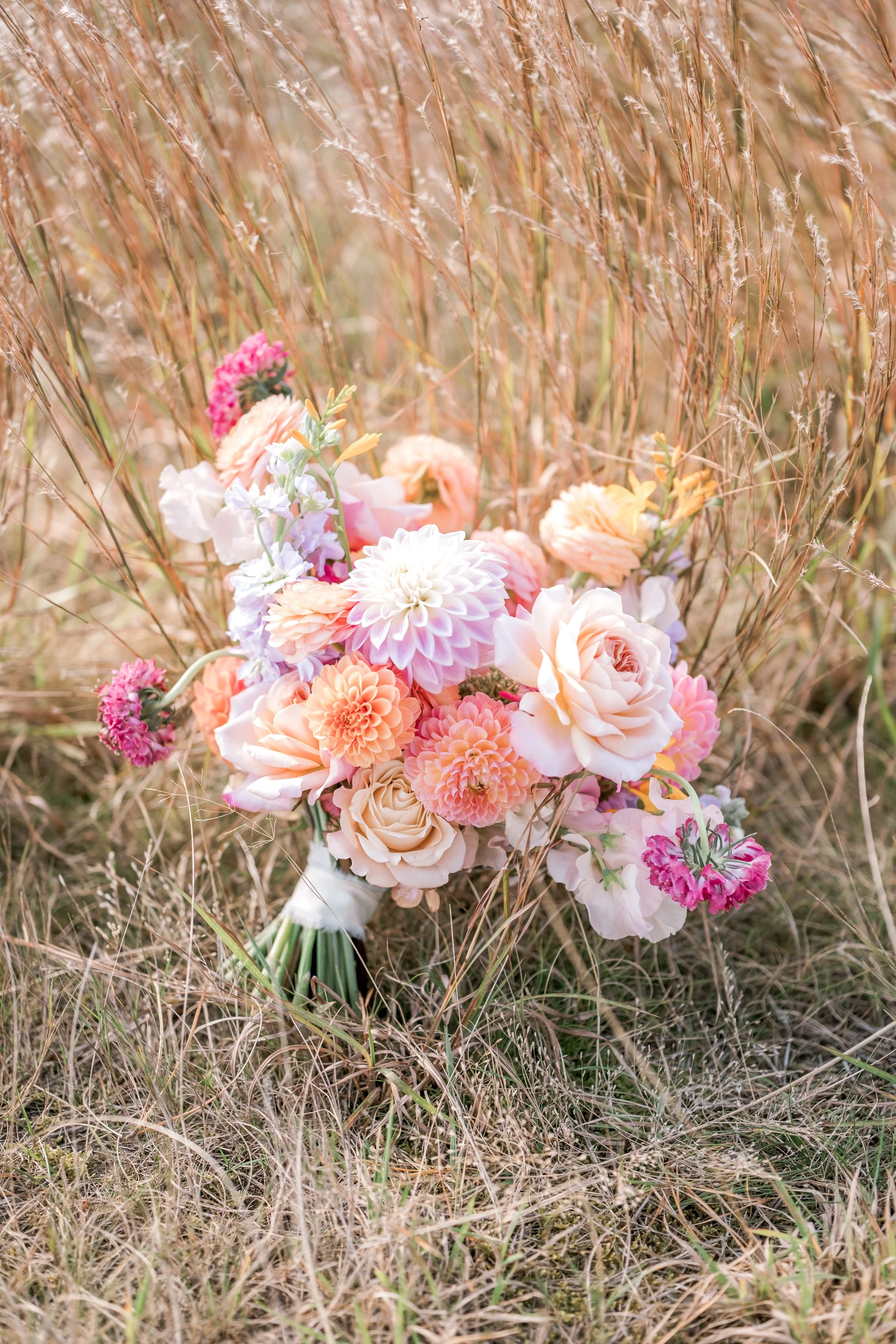 A bouquet of pink, white, and peach flowers resting on dry grass in a natural outdoor setting.