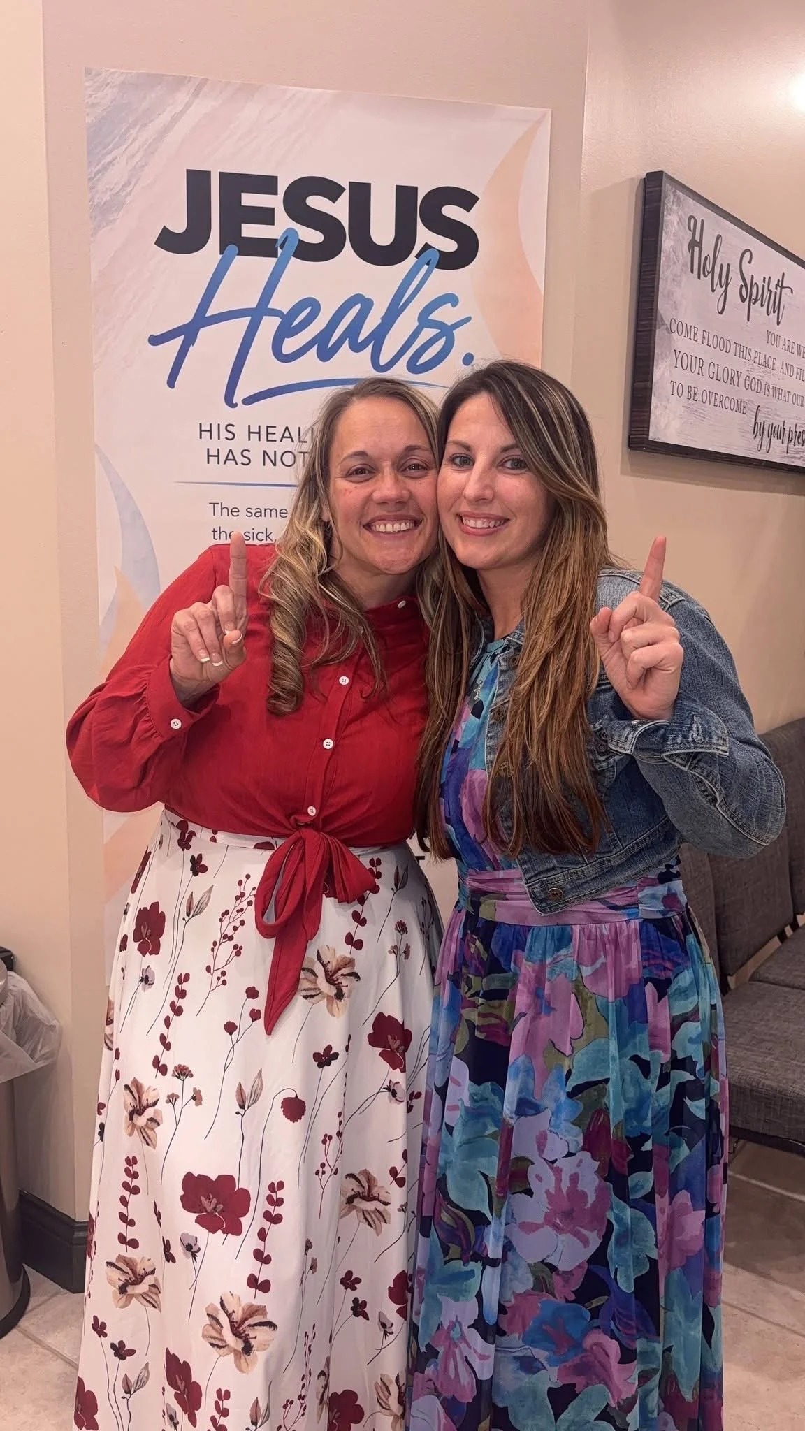 Two women smiling and pointing upward beside a “Jesus Heals” sign.