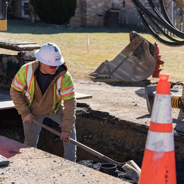 Construction worker in safety gear working on a road project, with an excavator bucket and safety cones nearby.
