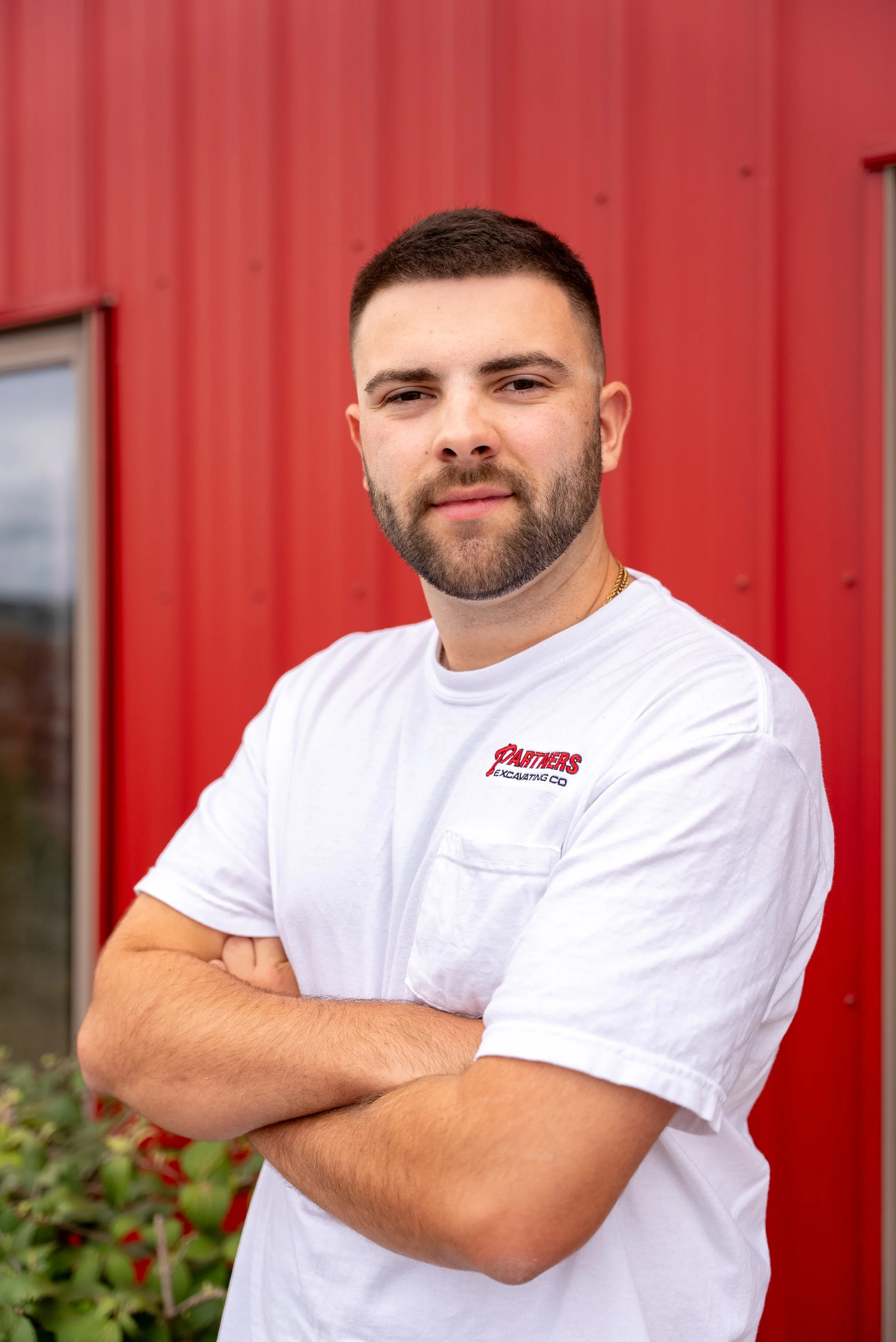 A young man with light skin and facial hair, wearing a white t-shirt with a logo, smiling with arms crossed, standing in front of a red building.