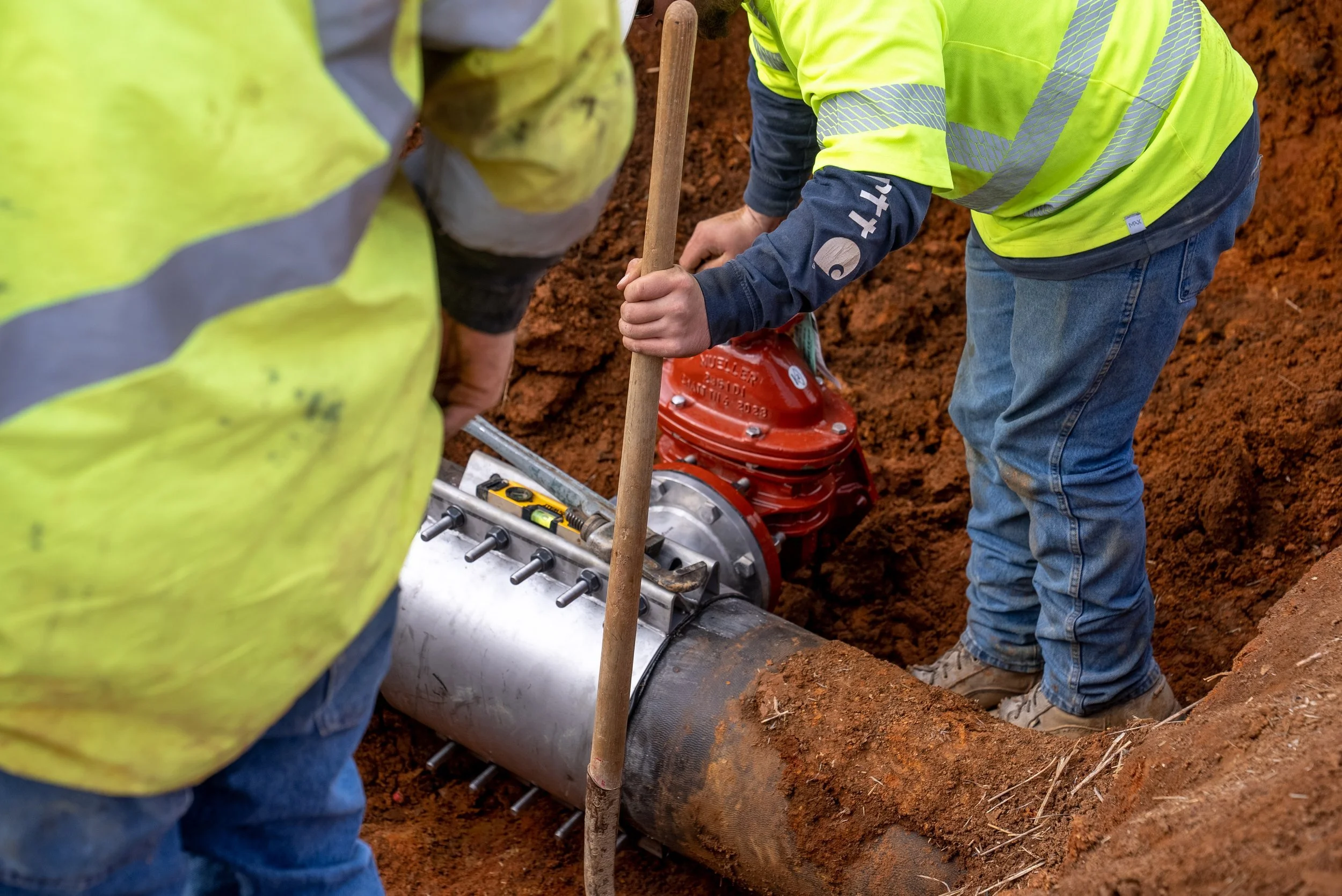 Workers in yellow safety vests installing or repairing a pipeline underground, using tools in a trench.