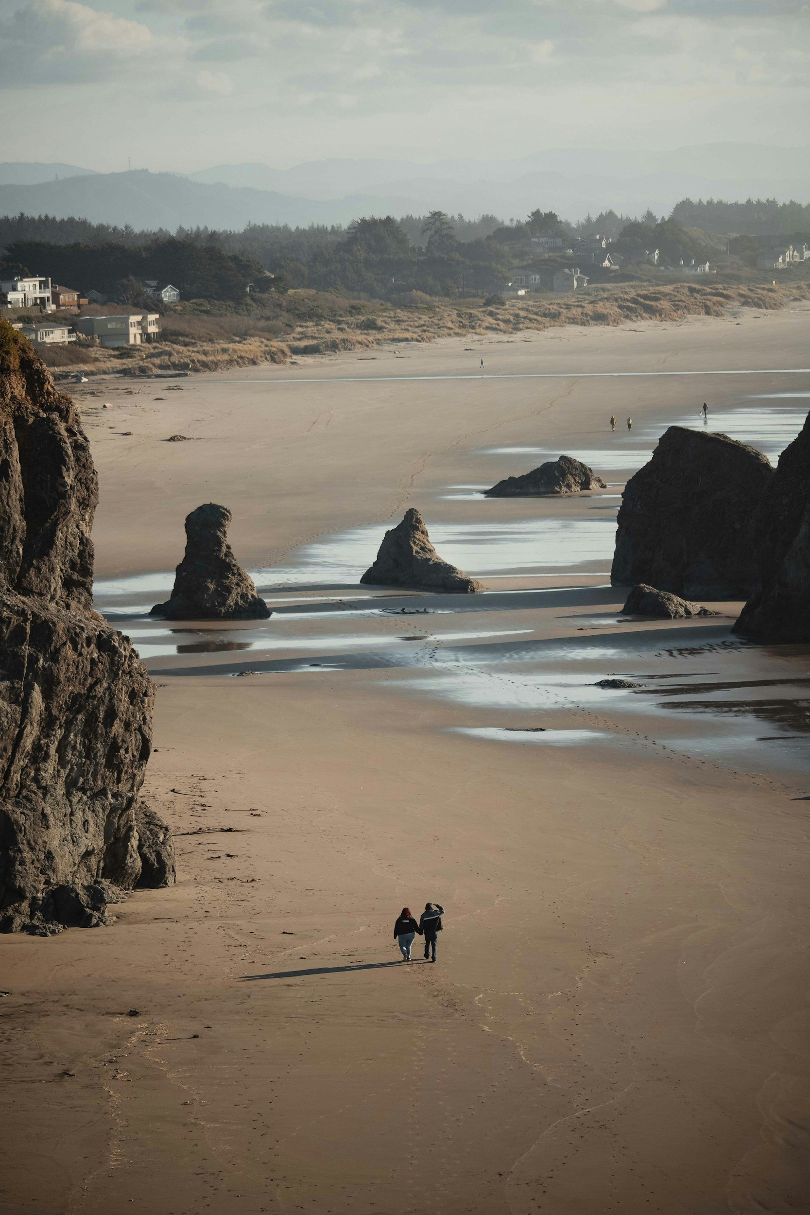 Couple walking on a sandy beach near large rocks with a scenic view of distant houses and hills in the background.
