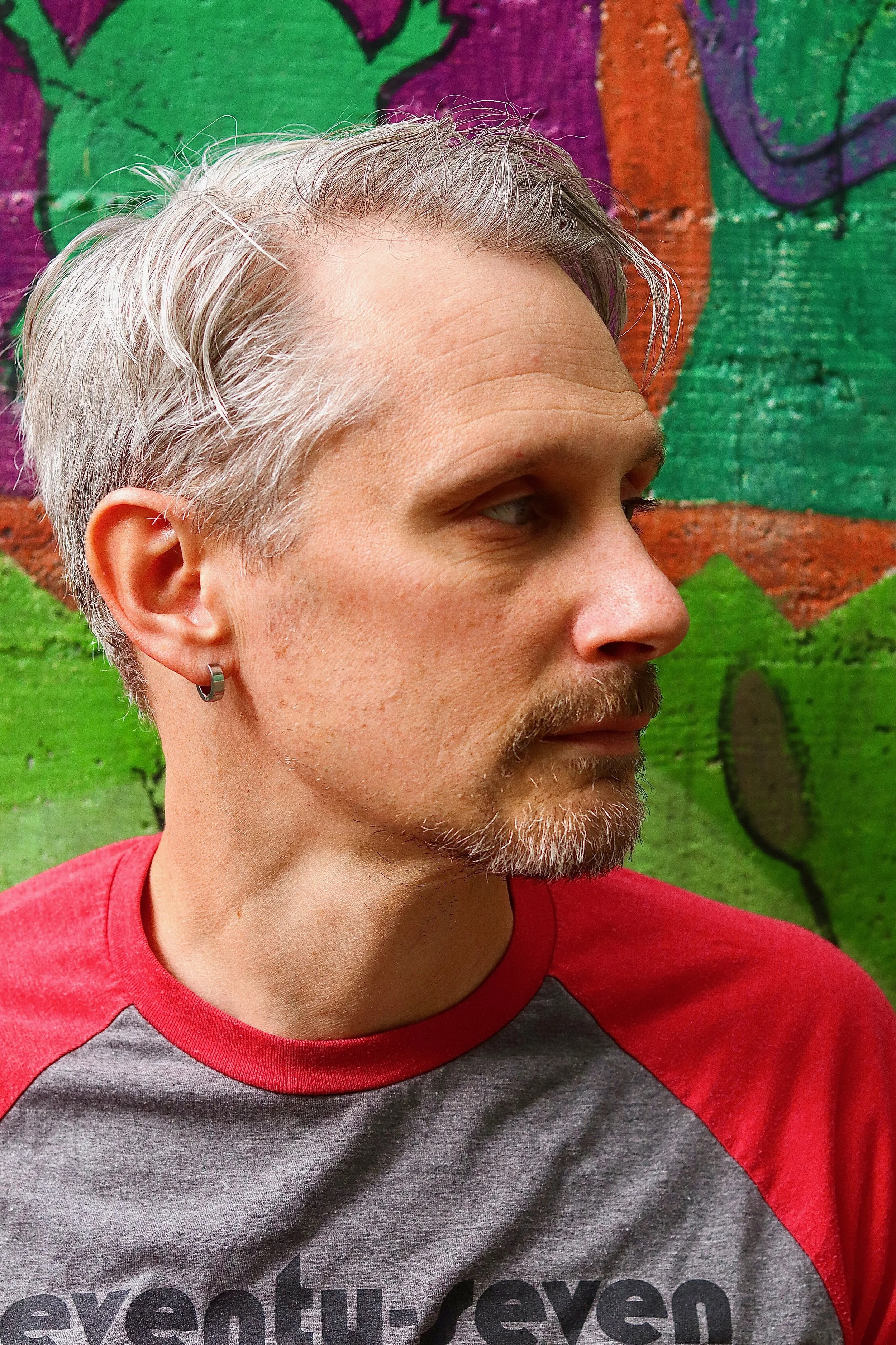 A young man with gray hair, a beard, and a hoop earring in his left ear looking to the right. He is wearing a gray and red shirt and standing in front of a colorful graffiti wall with green, purple, orange, and black designs.