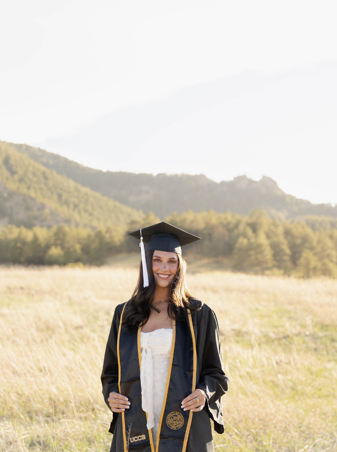 A young woman in a graduation gown and cap standing outdoors in a field with mountains and trees in the background, smiling.
