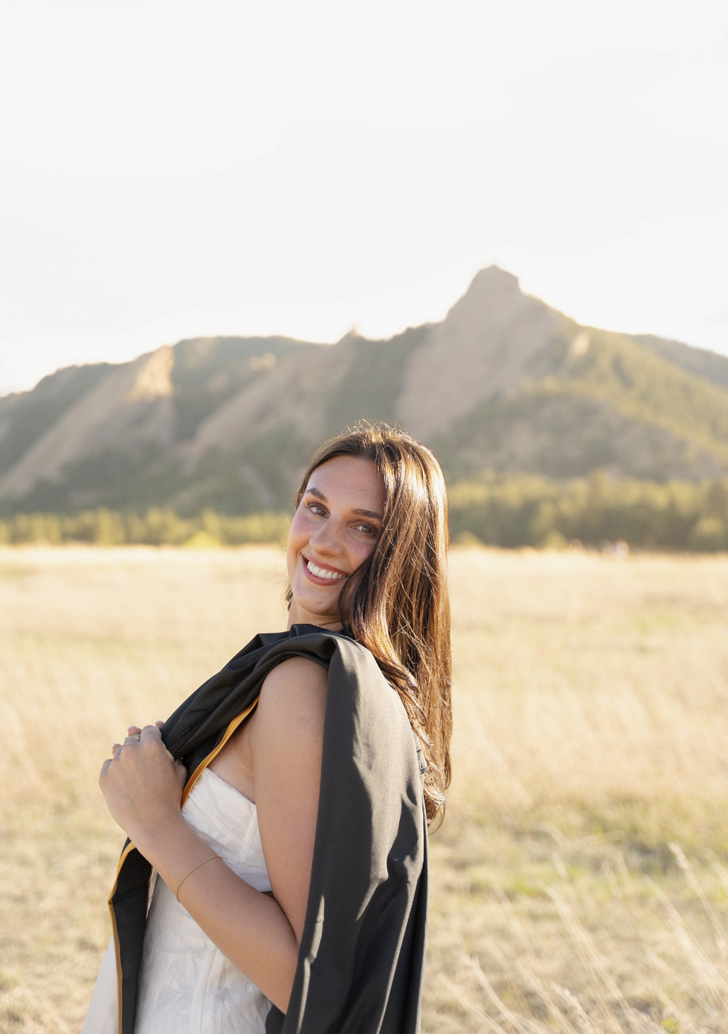A woman with long brown hair smiling at the camera, standing in a field with mountains in the background, wearing a white dress and holding a jacket over her shoulder.