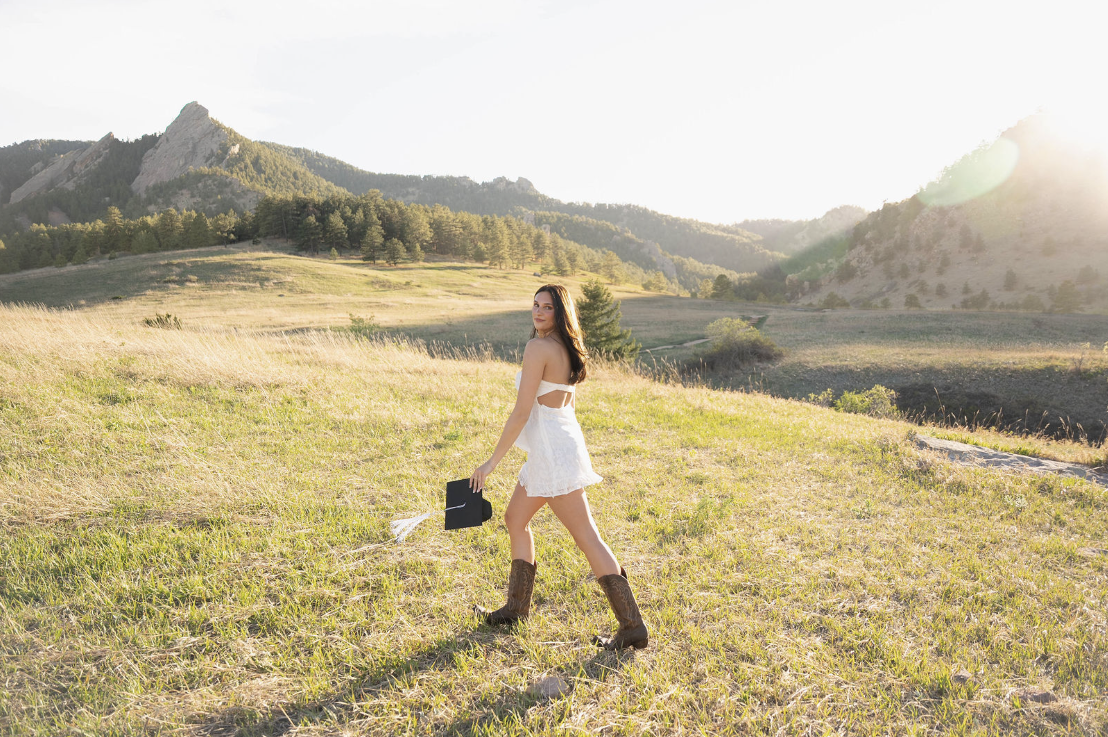 A woman in a white dress and cowboy boots walking through a grassy field with mountains and trees in the background, holding a black folder.