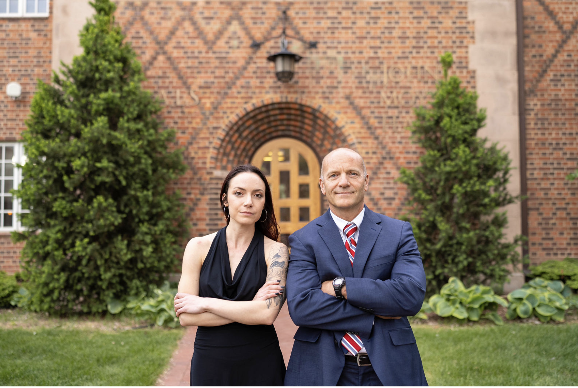 A man and a woman stand outside in front of a brick building with trees and bushes. The woman has long dark hair, tattoos on her arms, and wears a black sleeveless dress. The man is bald, wears a blue suit with a striped red, white, and blue tie, and