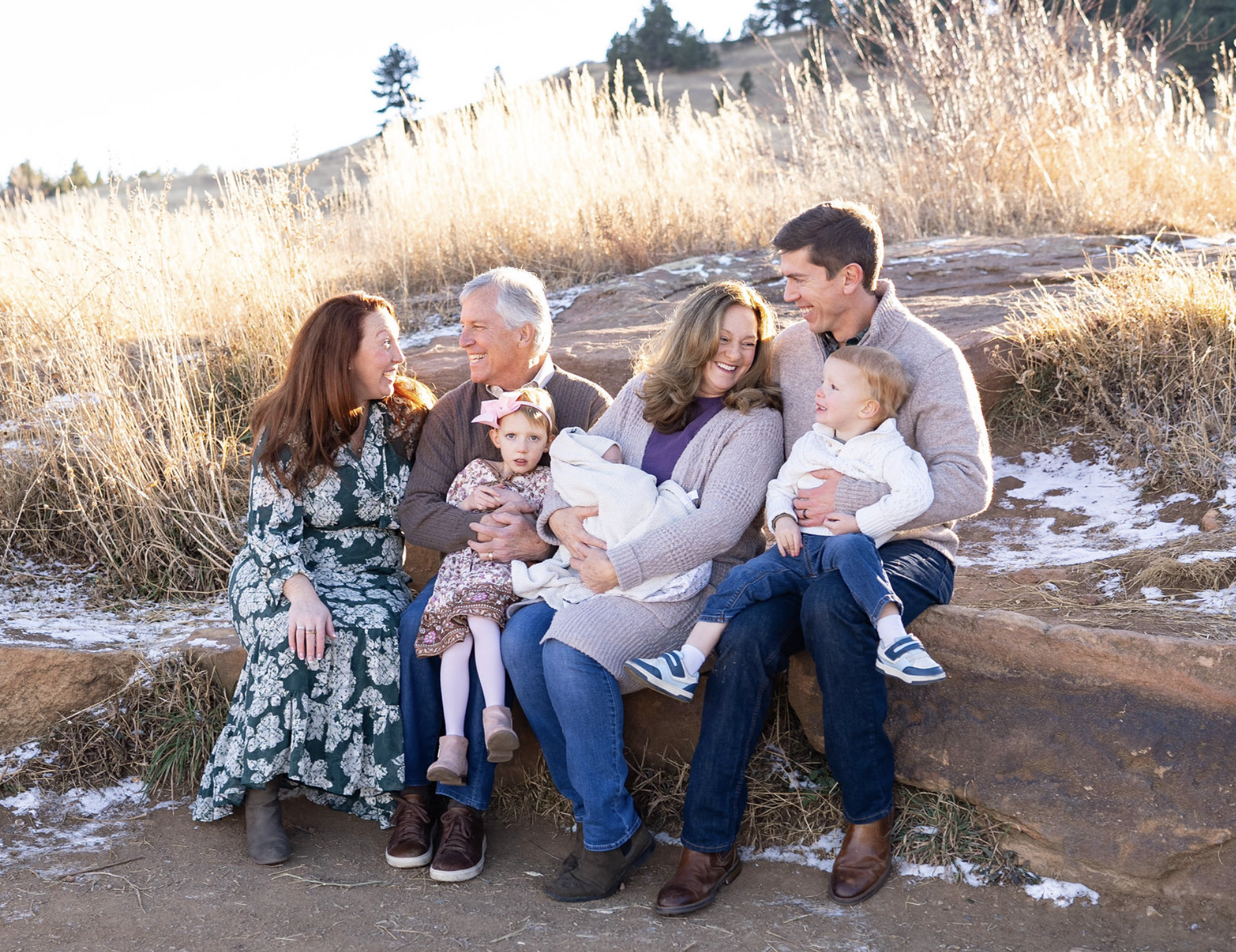 A multigenerational family of six sitting and standing outdoors on a rocky area, smiling and laughing together during daytime with golden grass and trees in the background, some snow on the ground.