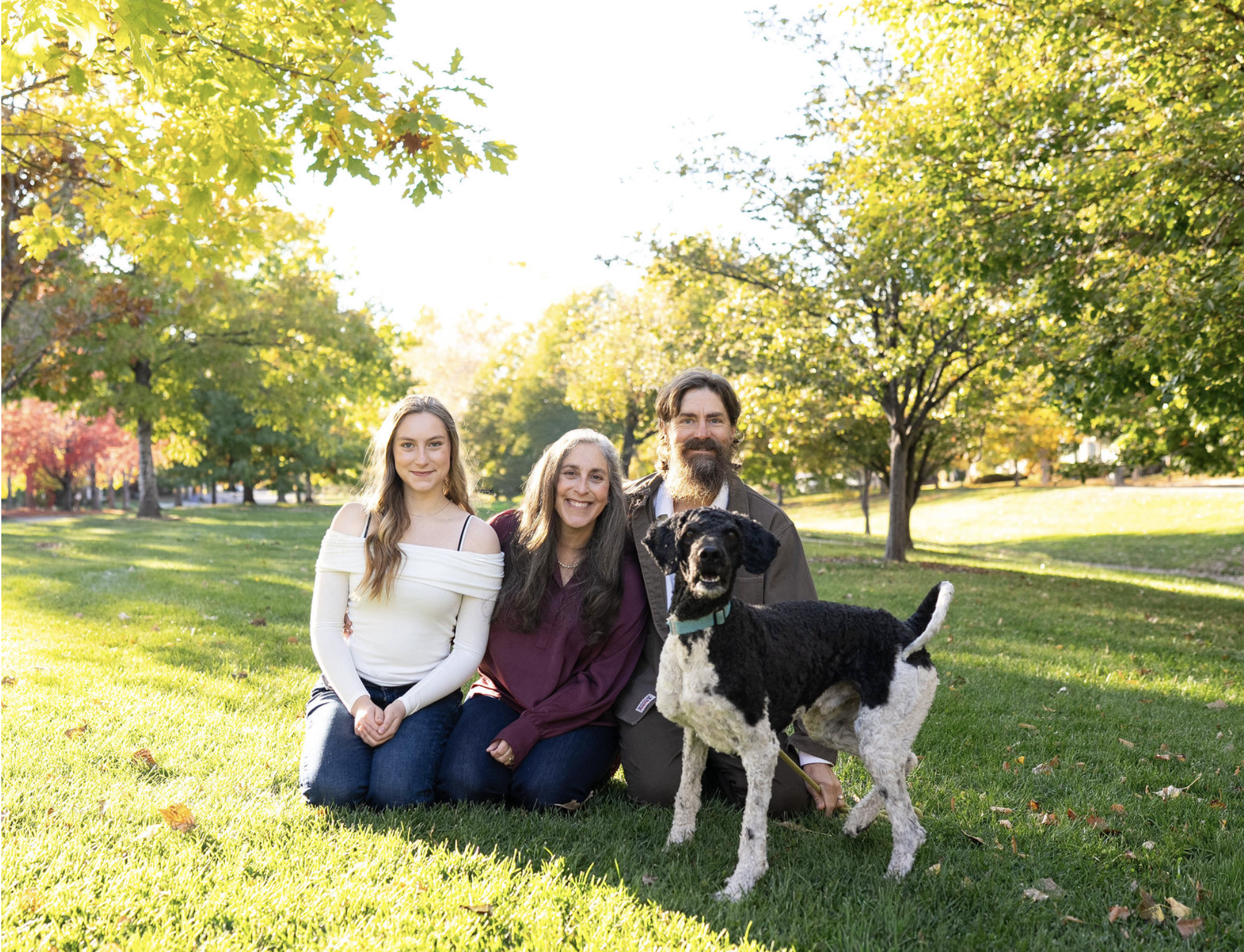 Three women and a man, along with a black and white dog, sitting on grass in a park with green and yellow autumn leaves on trees, under a bright sky.