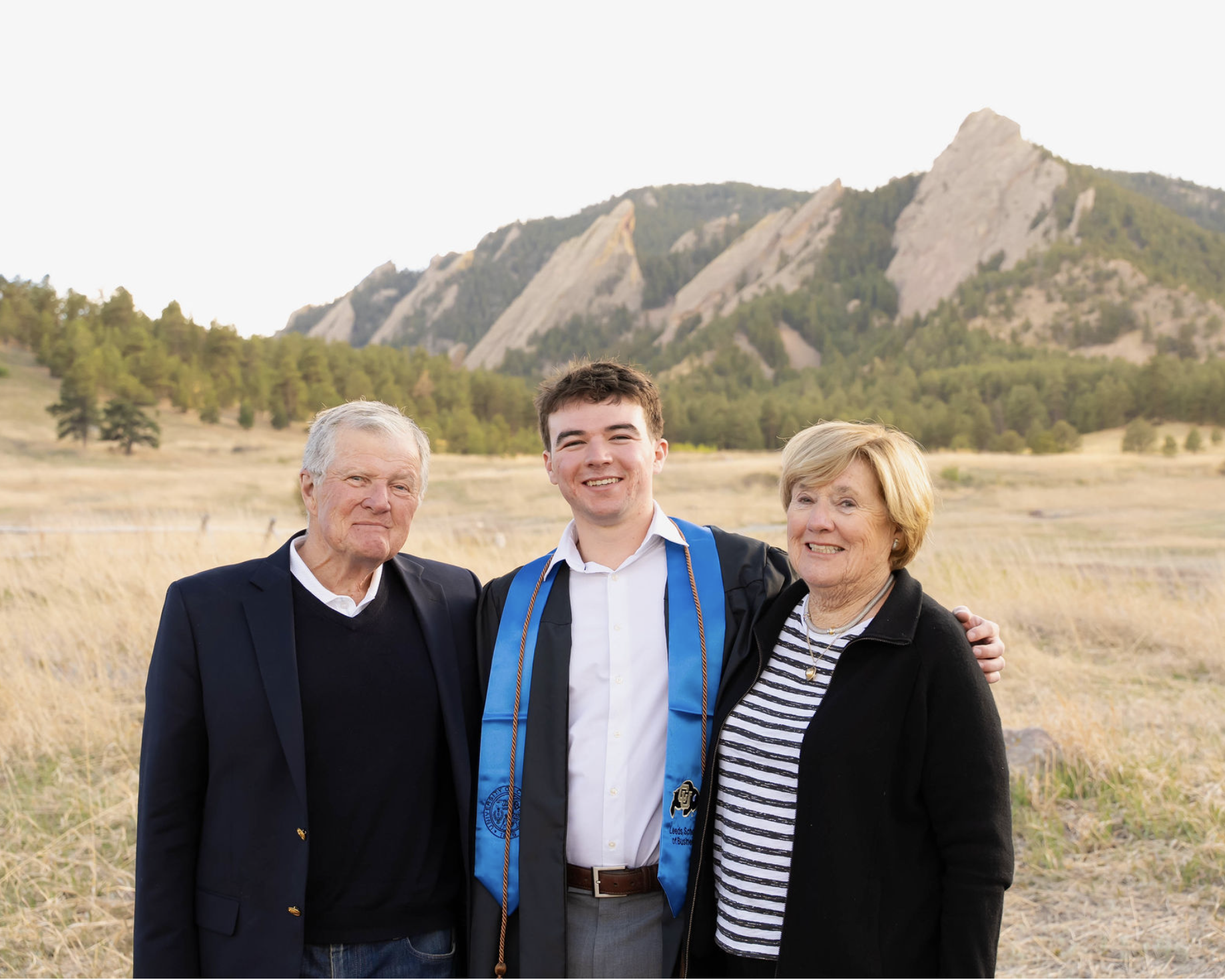 A young man in a graduation gown and cap standing between an older man and woman in a field with mountains in the background.