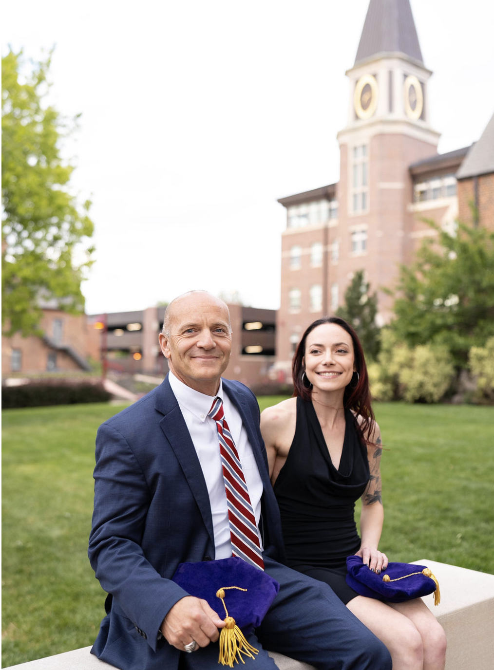 A man and woman sitting outdoors on a bench, holding graduation caps, with a castle-like building and trees in the background.