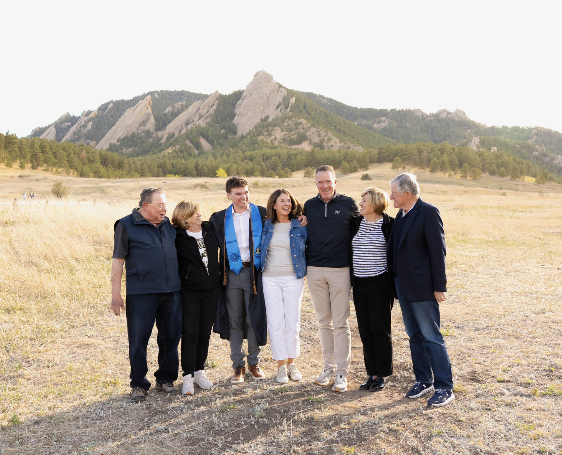 A group of seven people standing outdoors in a field with mountains in the background, smiling and hugging each other.