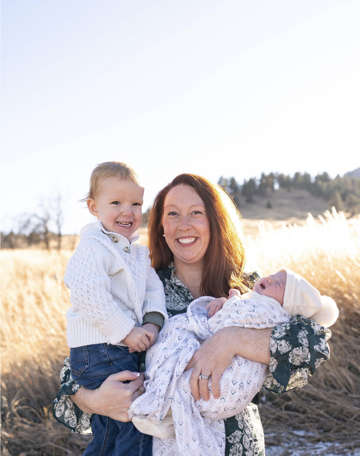 A woman holding a sleeping baby wrapped in a blanket, standing outdoors in a field during sunset, with a young child smiling beside her.