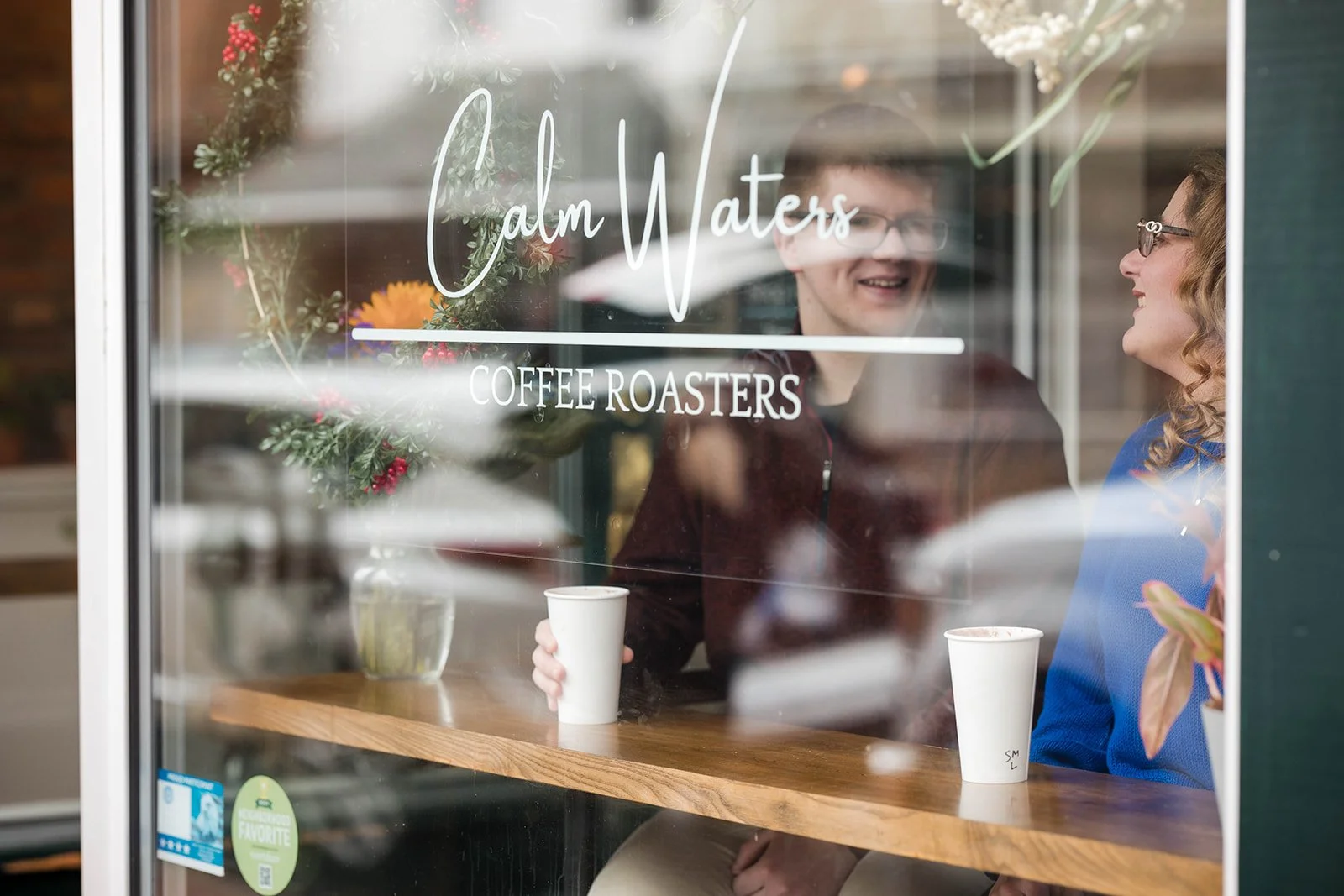 a couple sits in the window of Calm Waters Coffee Roasters smiling and drinking coffee