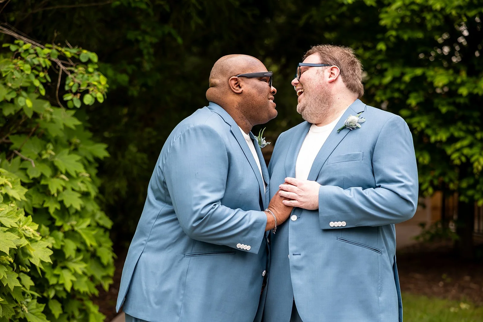 Two grooms, in matching light blue suits, laughing with each other in front of foliage near McCloone's Boat House in West Orange, NJ.