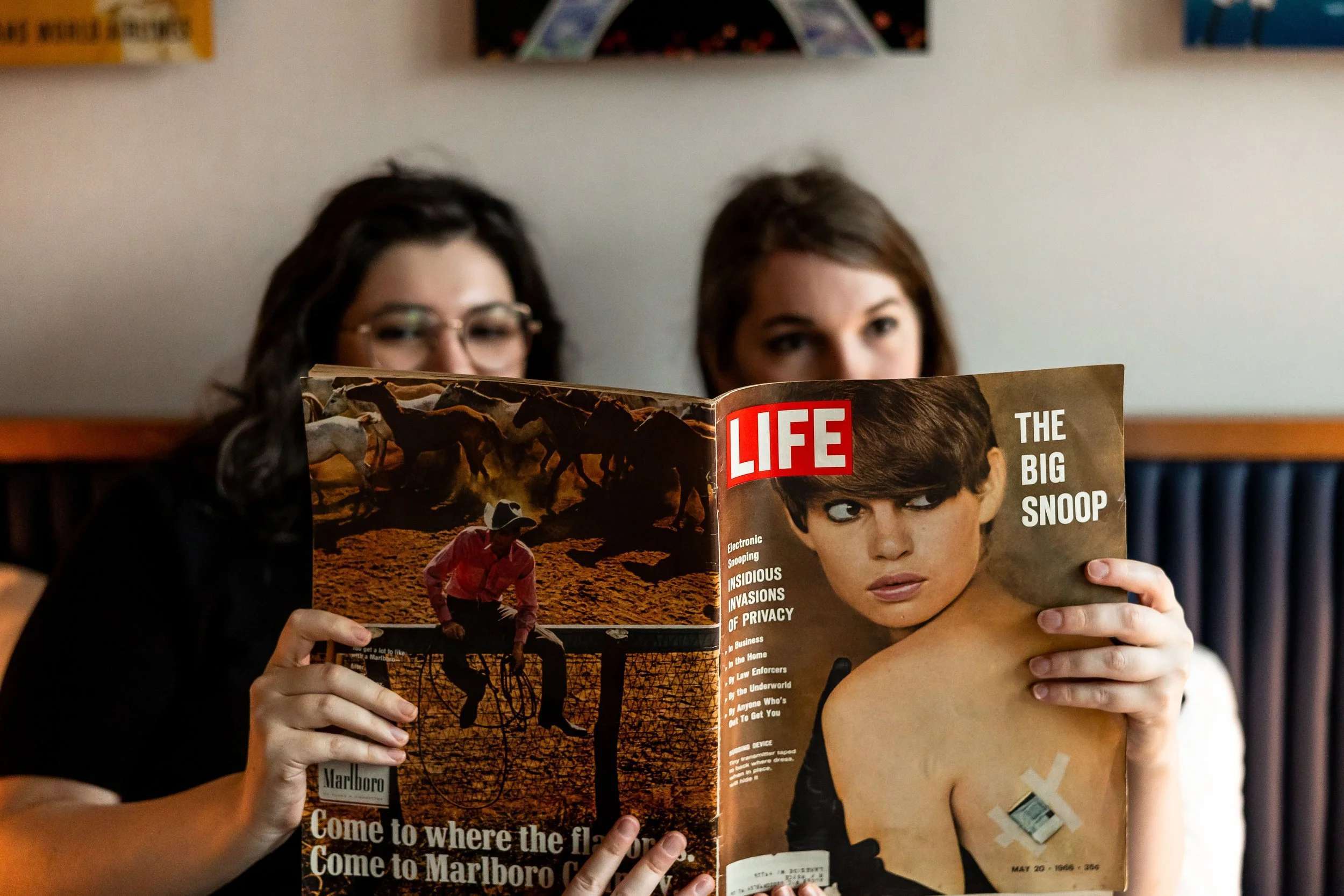 A couple reading a vintage LIFE magazine while laying in a bed at the TWA Hotel at JFK Airport in New York City. the magazine is covering most of their faces, but you can see their eyes.