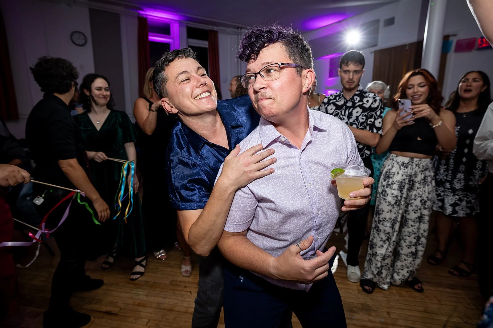 Two marriers dancing together at their venue Pig Iron Theater in Philadelphia, PA. One has a drink in their hand, the other is behind them, and they are looking at each other.