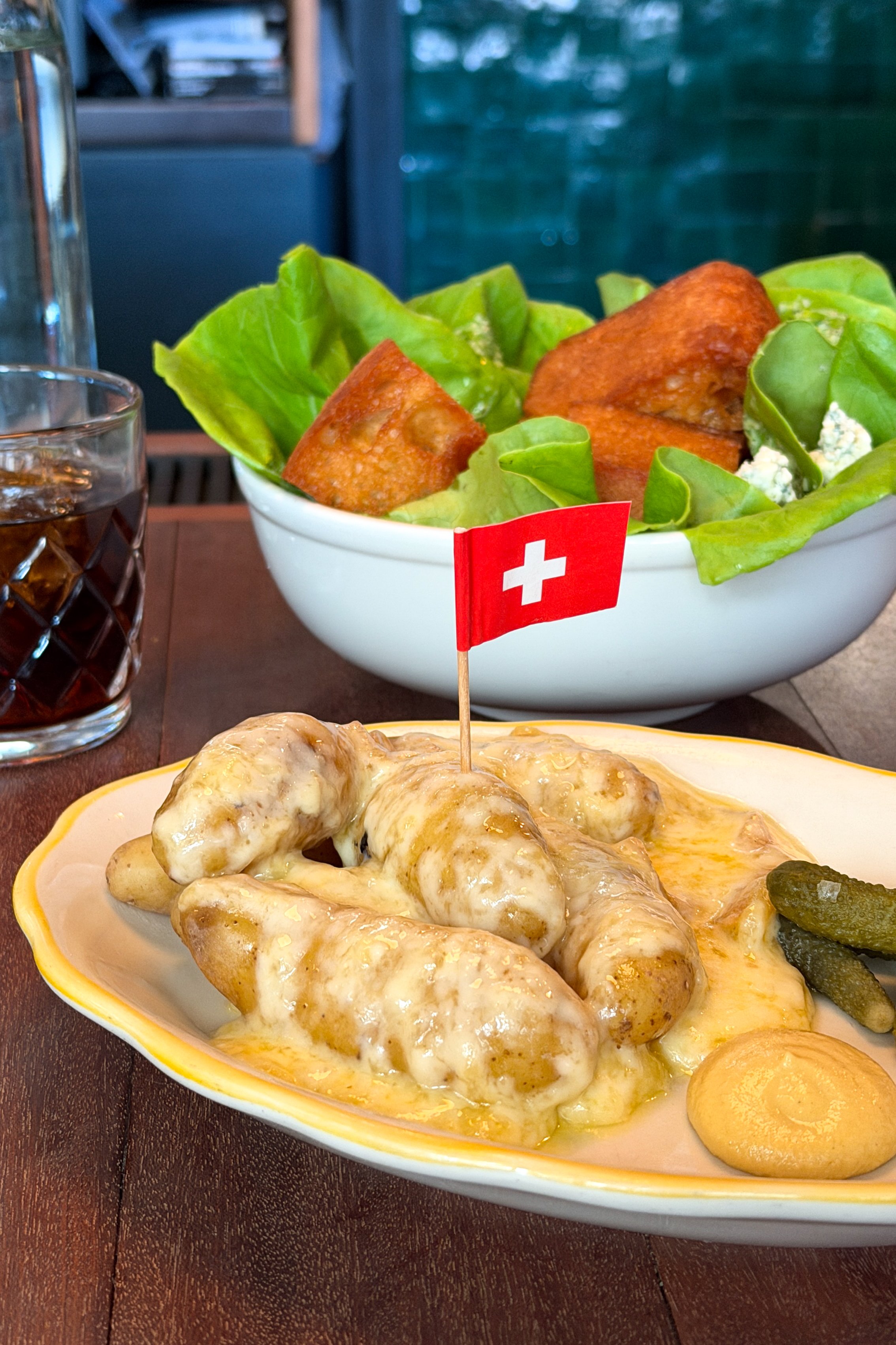 The raclette at The Bull and Beggar in Asheville, potatoes draped in melted cheese with a Swiss flag pick, served alongside the B&B Salad