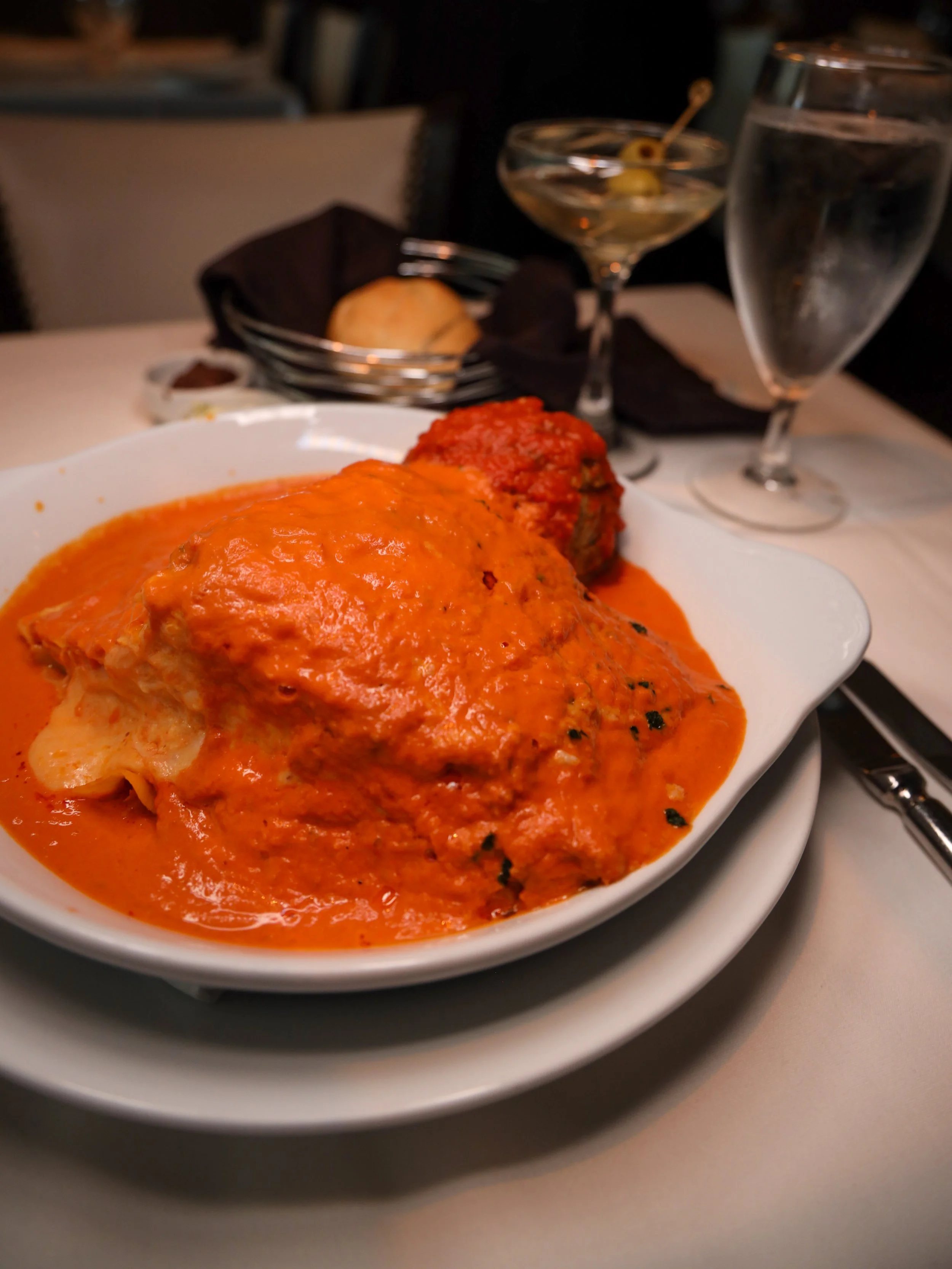 The Legendary Lasagna alla Vodka at Ristorante Paoletti, topped with a Wagyu beef and ricotta meatball, served alongside a bread basket and a martini.