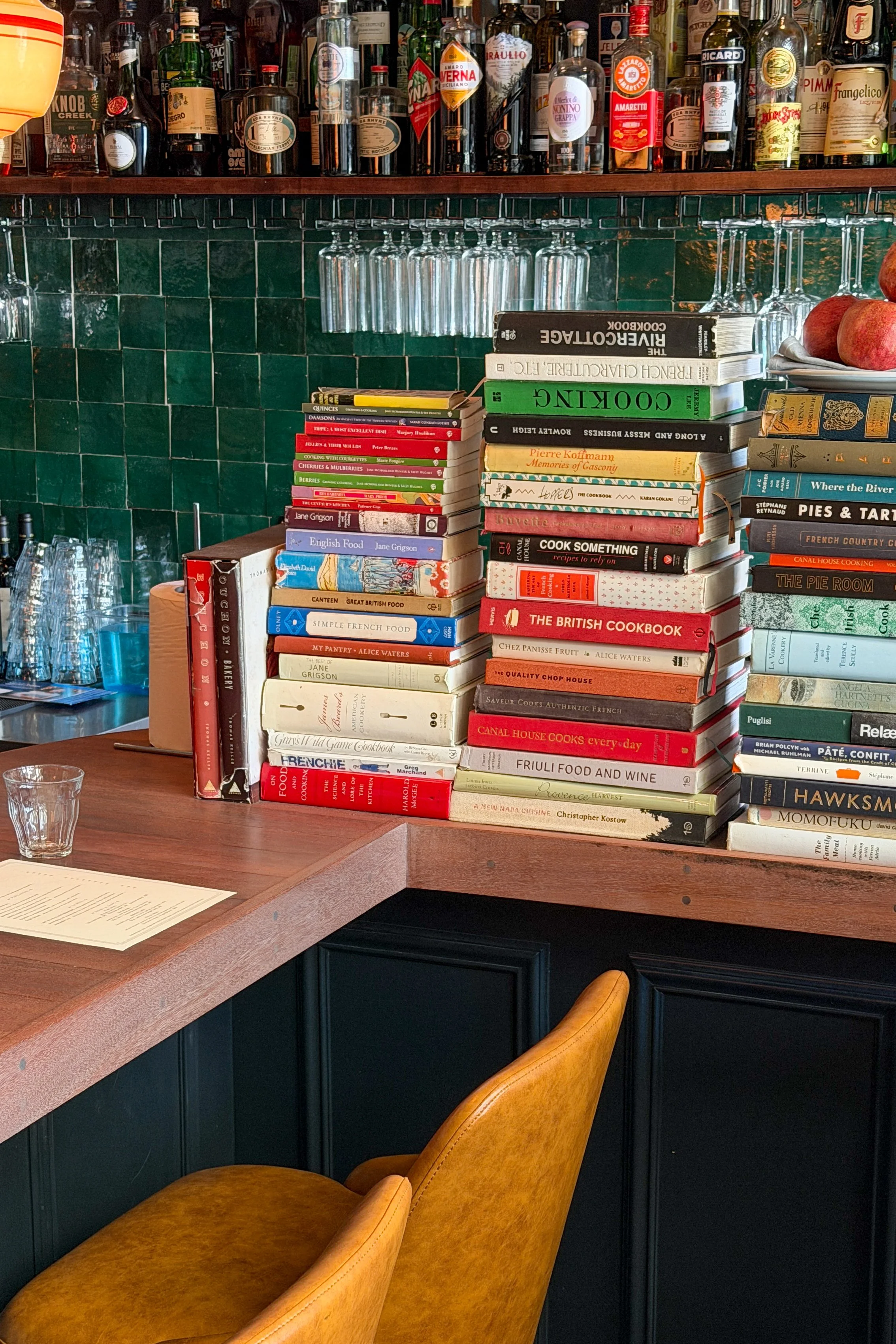 The bar counter at The Bull and Beggar in Asheville, stacked high with cookbooks beneath hanging wine glasses and yellow leather stools.