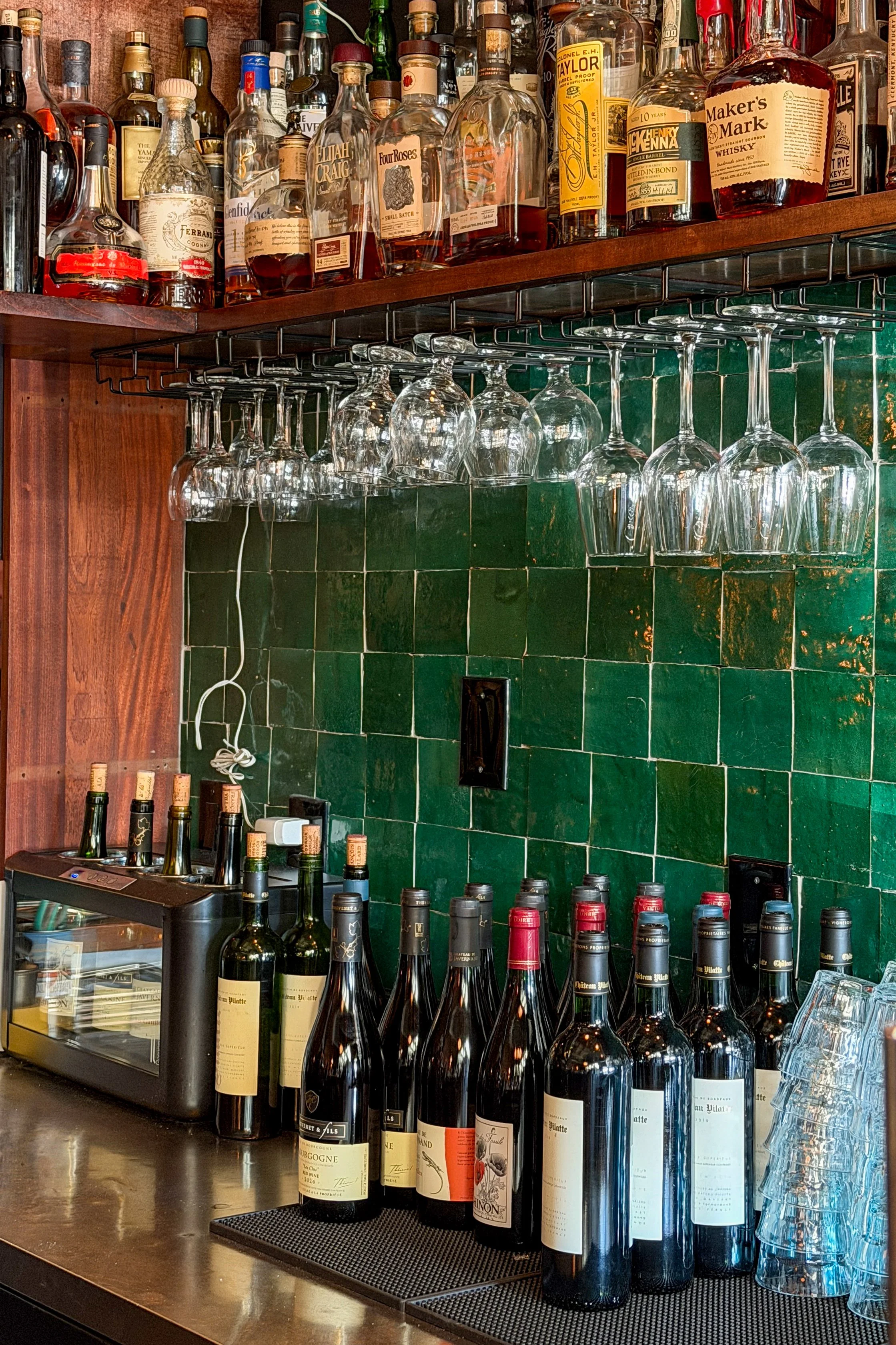 The bar at The Bull and Beggar in Asheville, lined with bourbon and cognac bottles above a green tile backsplash and hanging wine glasses.