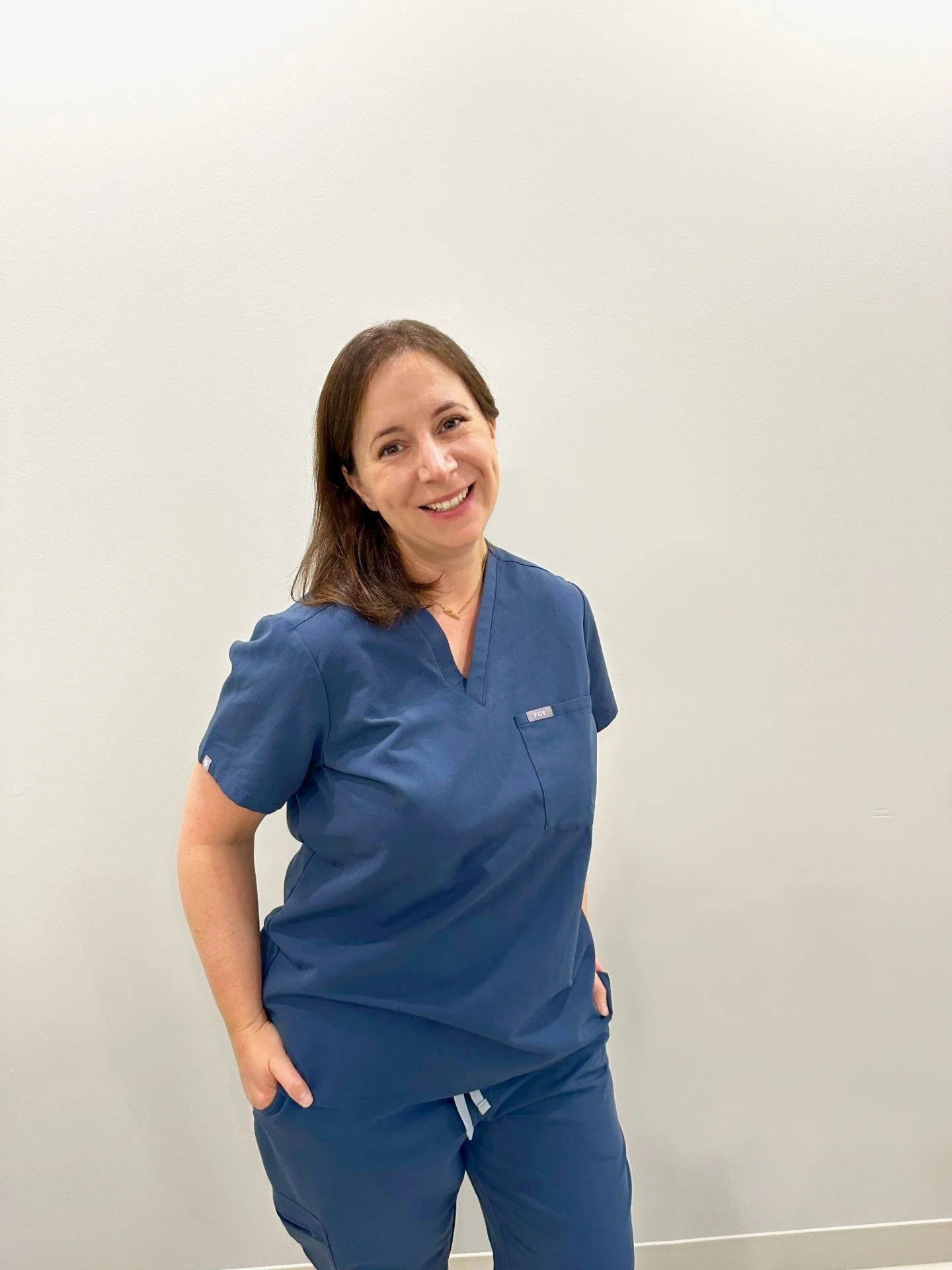 A woman in blue medical scrubs standing against a plain white wall, smiling at the camera.