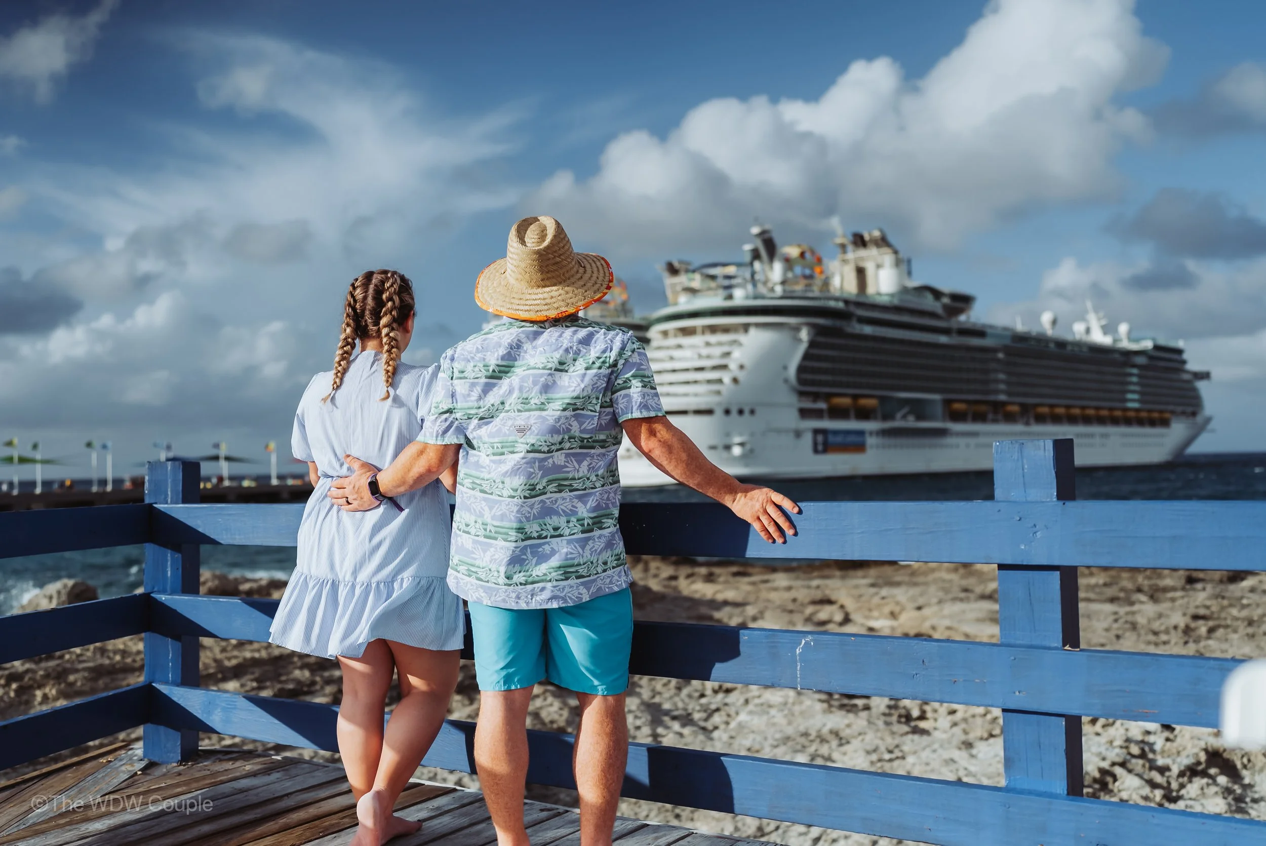 Older man and young girl with braided hair standing on a wooden pier, watching a large cruise ship on the water.