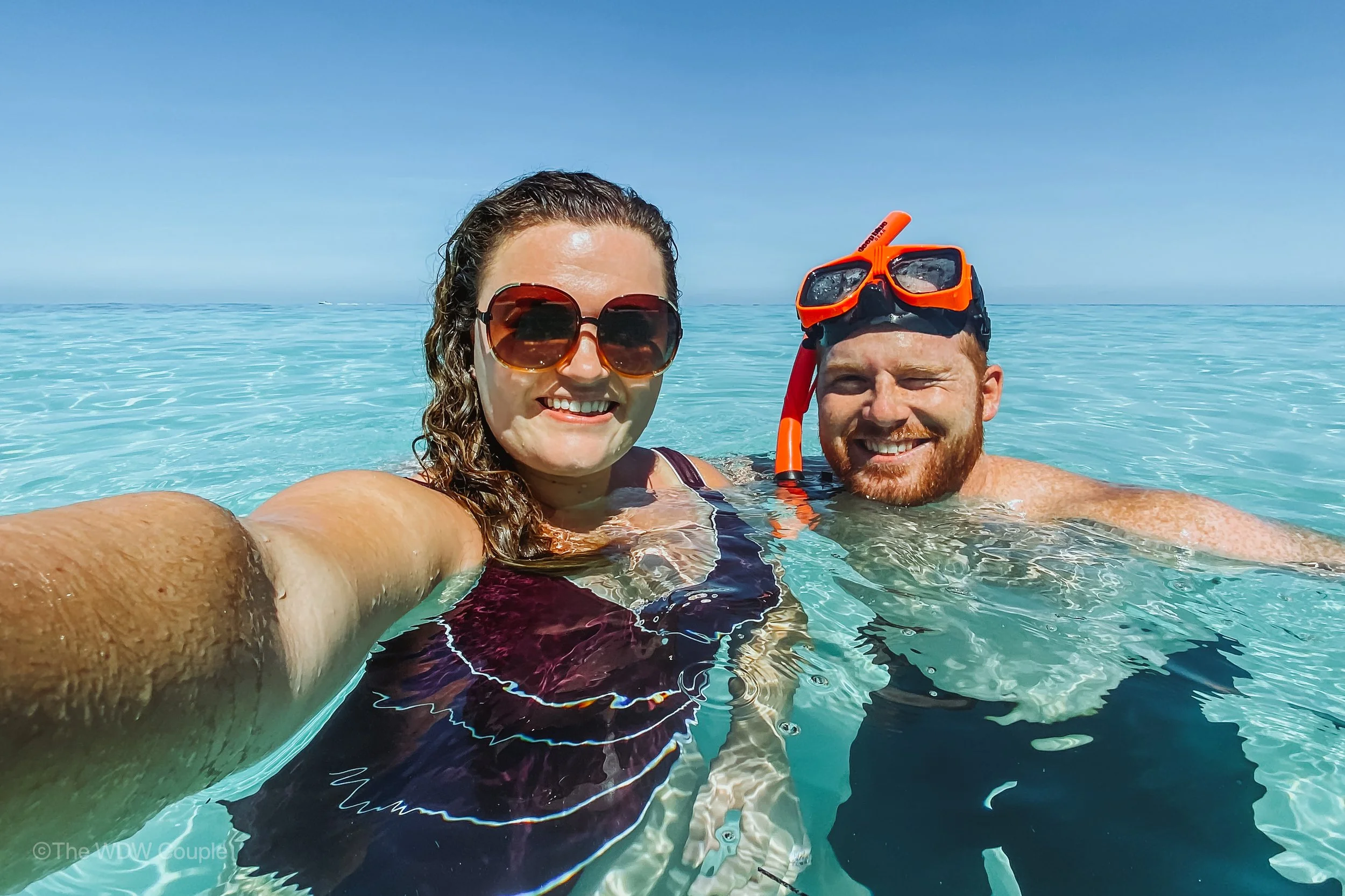 A smiling woman and man taking a selfie in the ocean. The woman is wearing sunglasses and a swimsuit, and the man has an orange snorkeling mask on his forehead. They are in clear, calm water with a blue sky above.